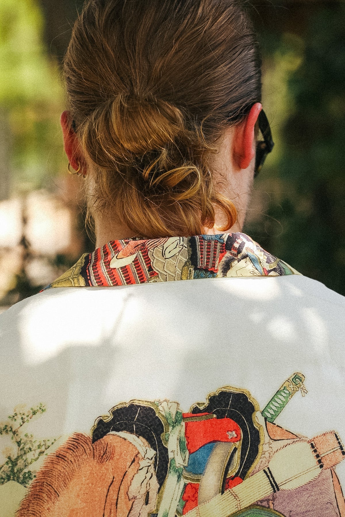A person viewed from behind wearing a white jacket with a large colorful graphic print on the back and a patterned collar, set against a blurred outdoor background.