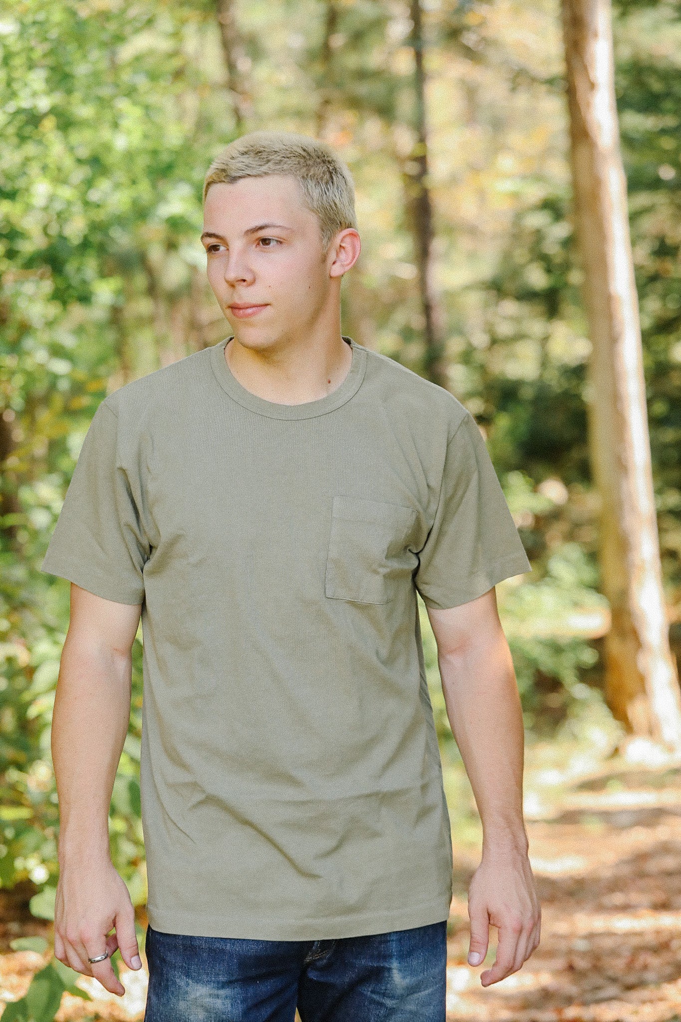 Young man outdoors wearing olive green pocket t-shirt and blue jeans in forest setting