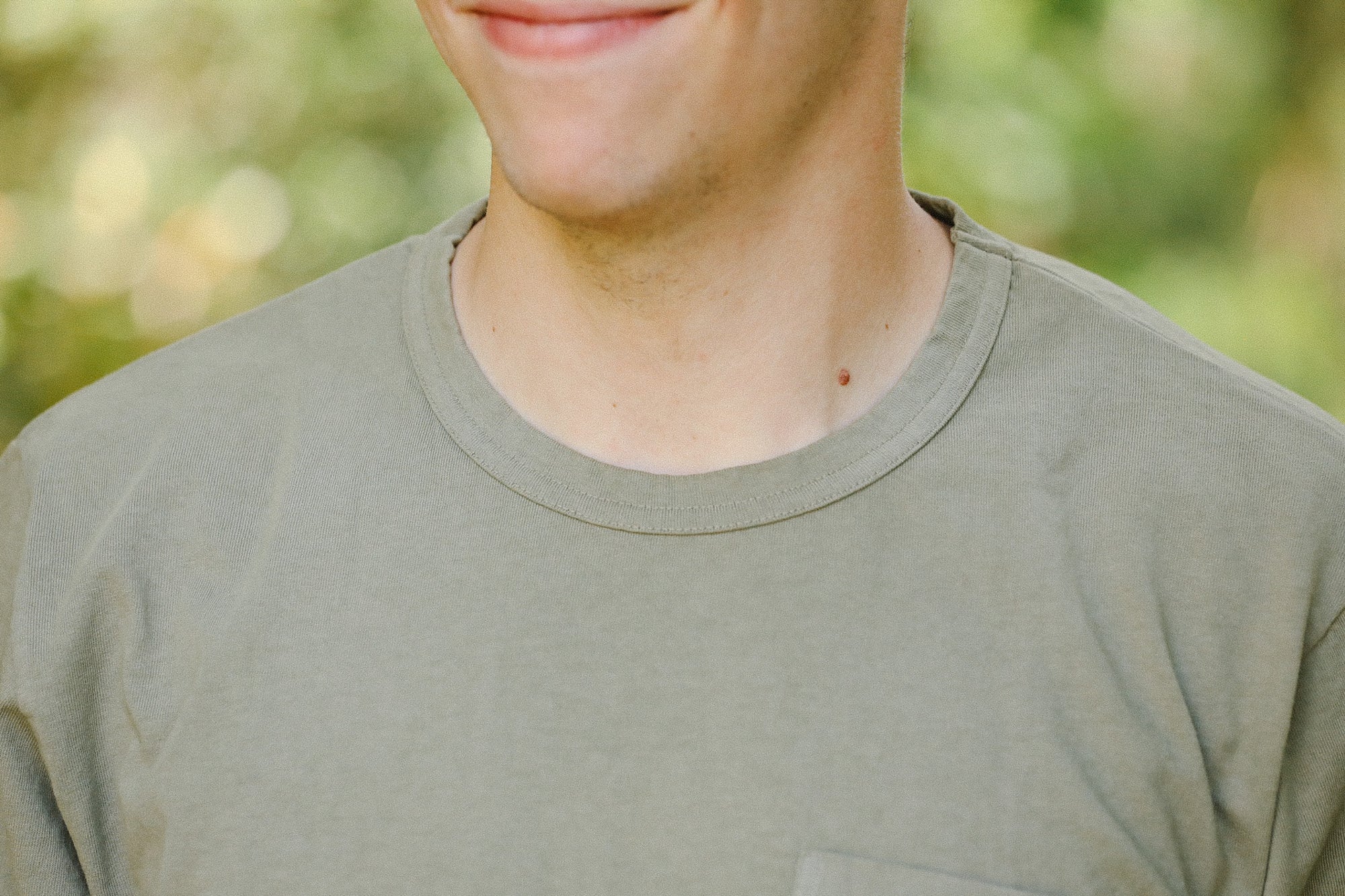 Close-up of a man wearing a light olive green t-shirt outdoors with blurred greenery background
