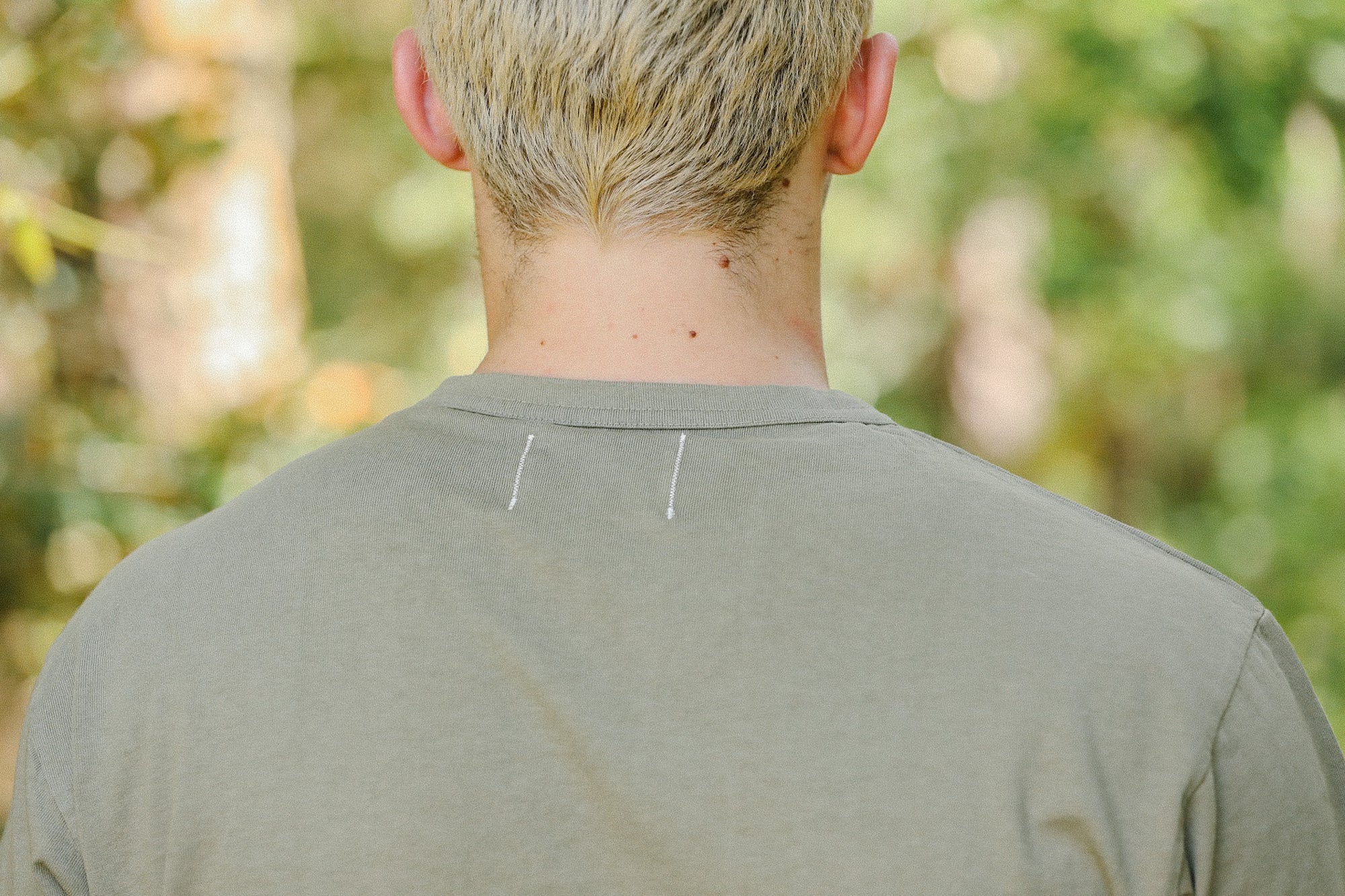 Back view of a person with short blonde hair wearing an olive green shirt outdoors with blurred greenery
