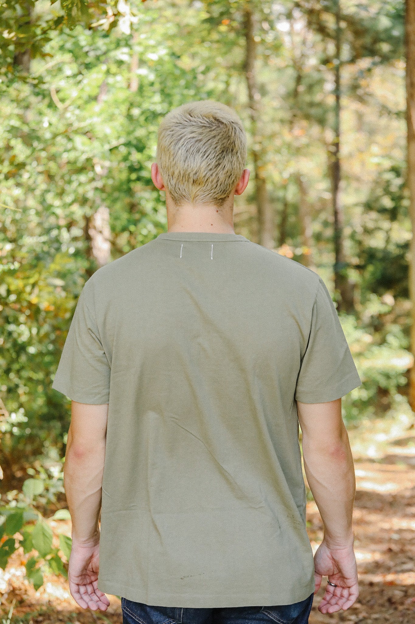 Man with blonde hair wearing an olive green t-shirt standing in a forest trail with trees and foliage