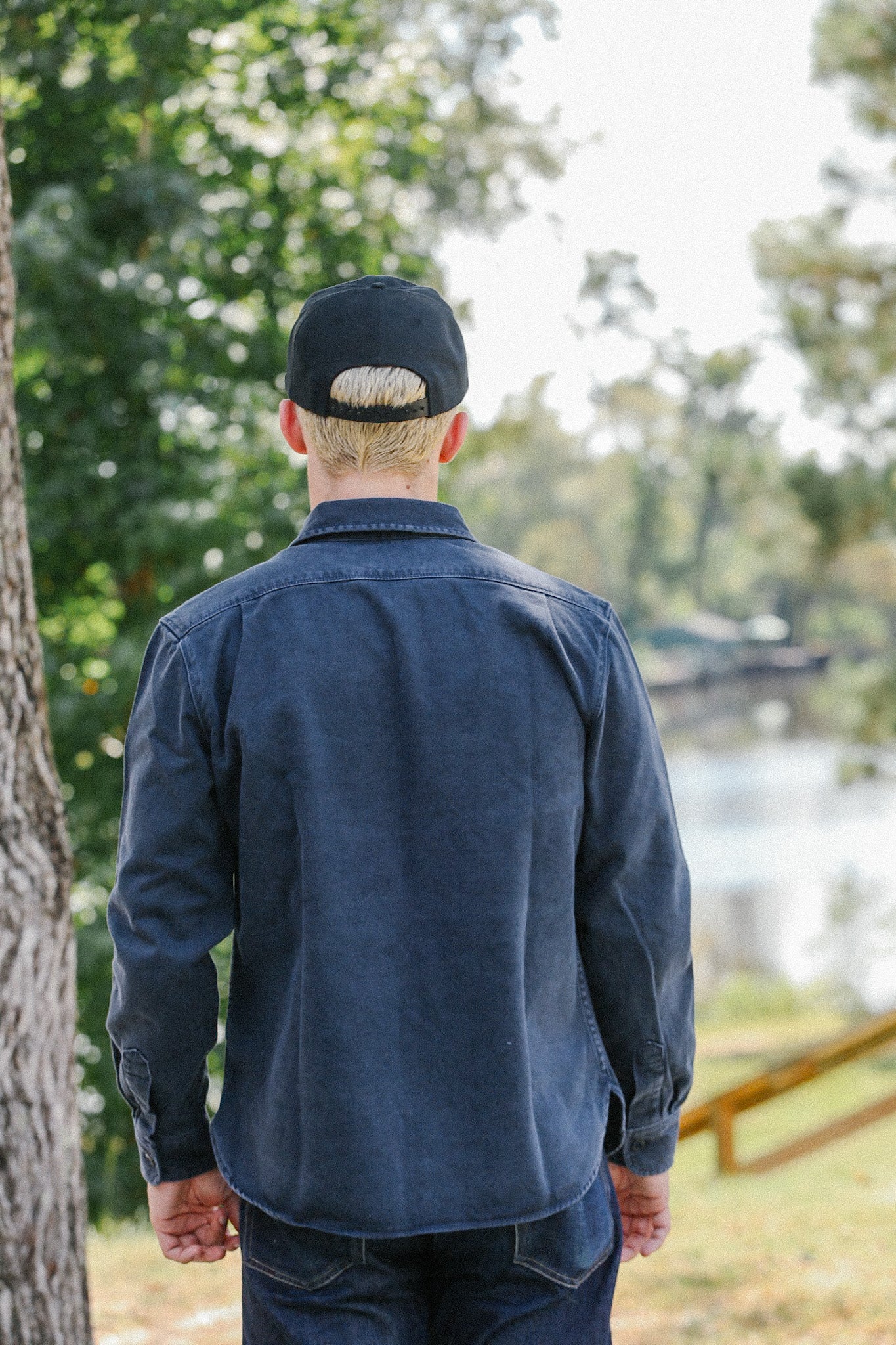 Man wearing dark blue jacket and black cap standing outdoors near tree and pond