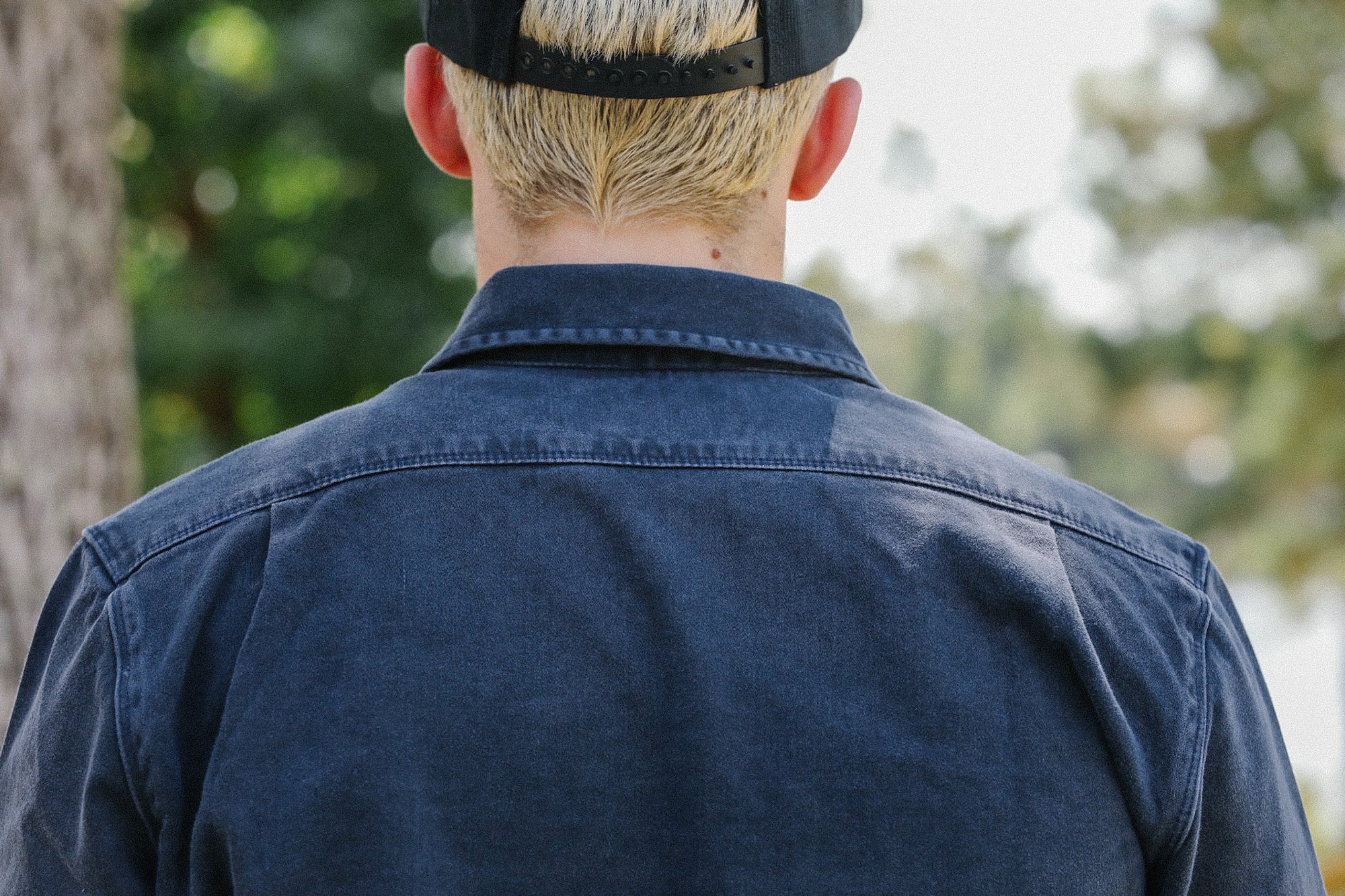 Back view of person wearing dark indigo work shirt and black snapback cap outdoors with blurred trees