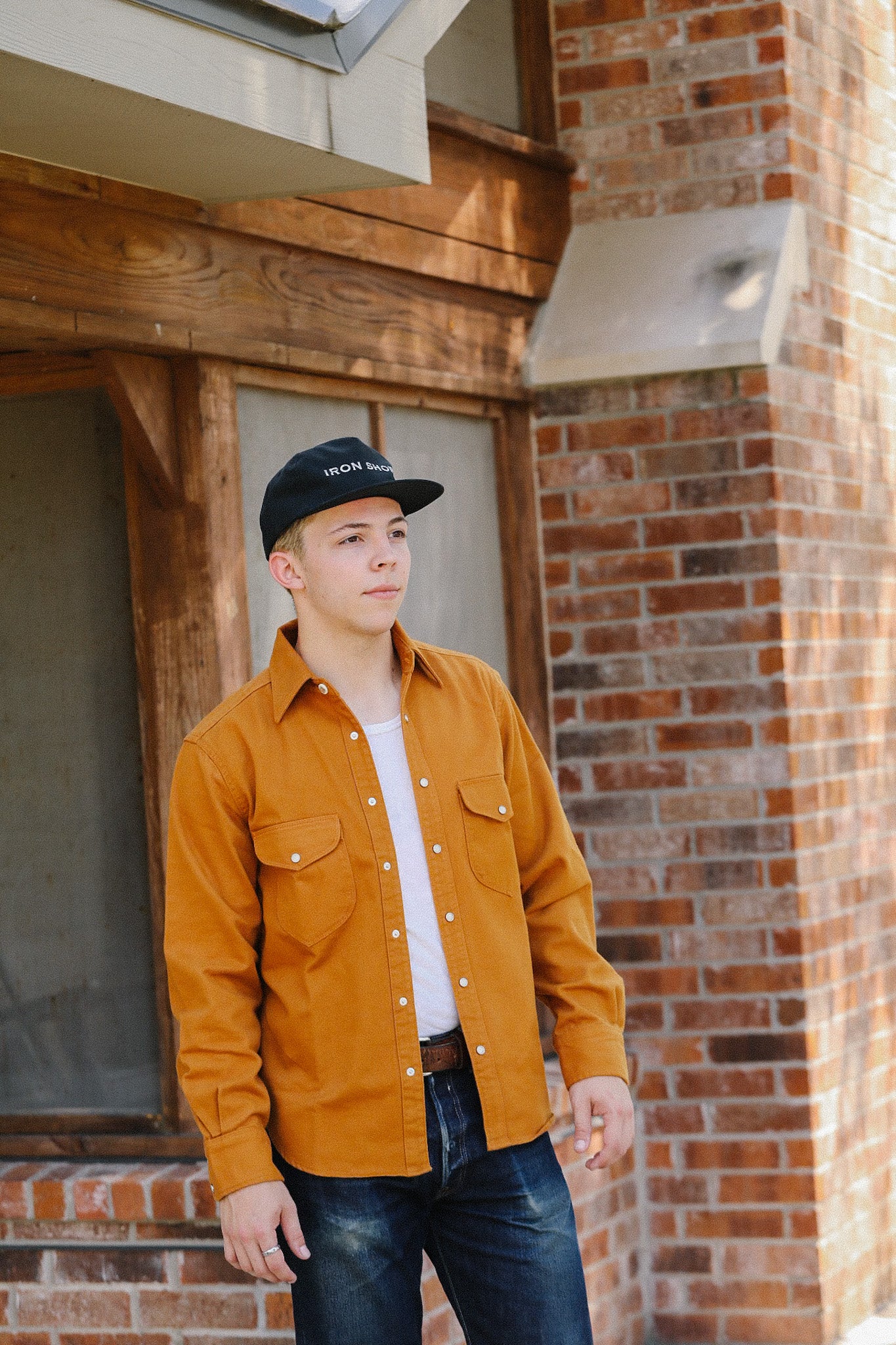 Young man wearing mustard work shirt, black Iron Shop Provisions cap, and jeans against brick and wood building