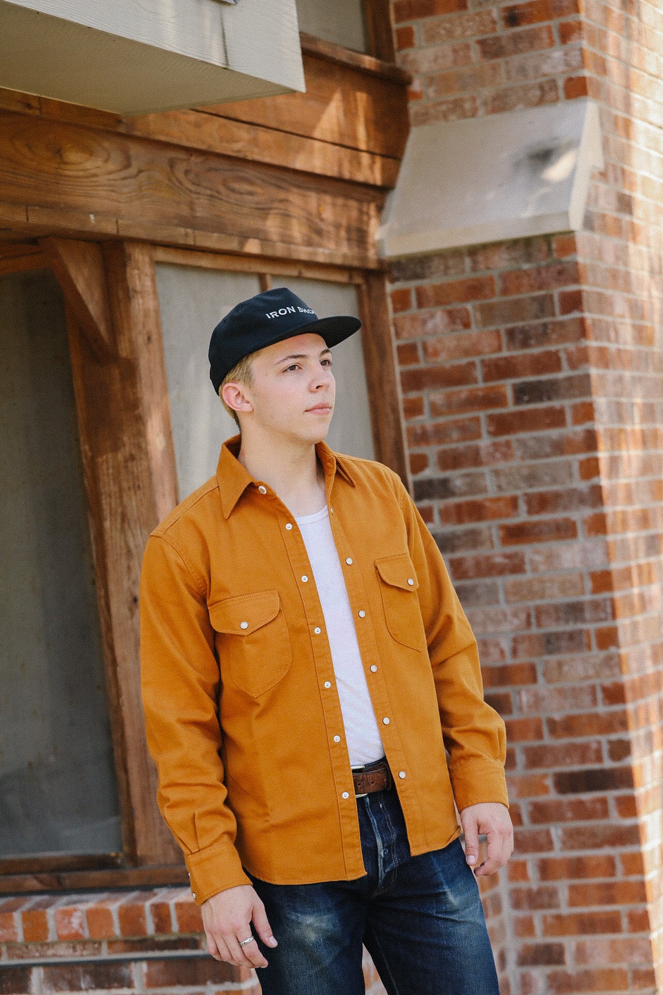 Young man wearing mustard shirt and Iron Shop Provisions cap standing by brick and wood building