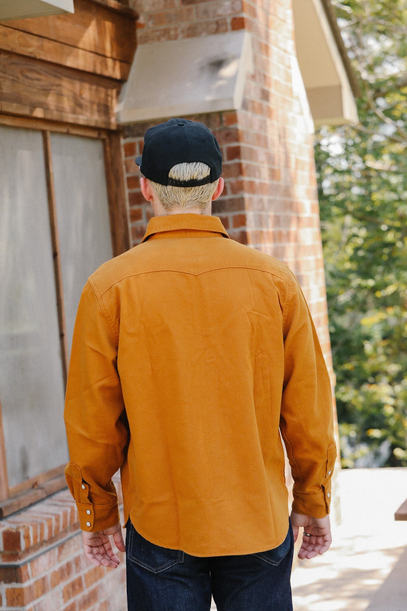 Man wearing a mustard work shirt and black cap standing outside a brick building, back view