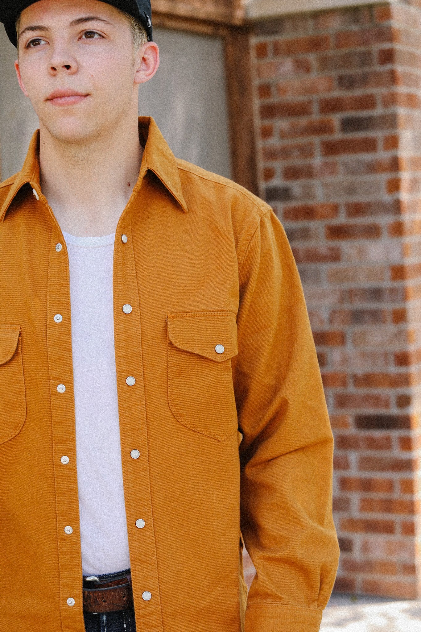 Young man wearing a mustard yellow work shirt and white undershirt standing in front of a brick wall