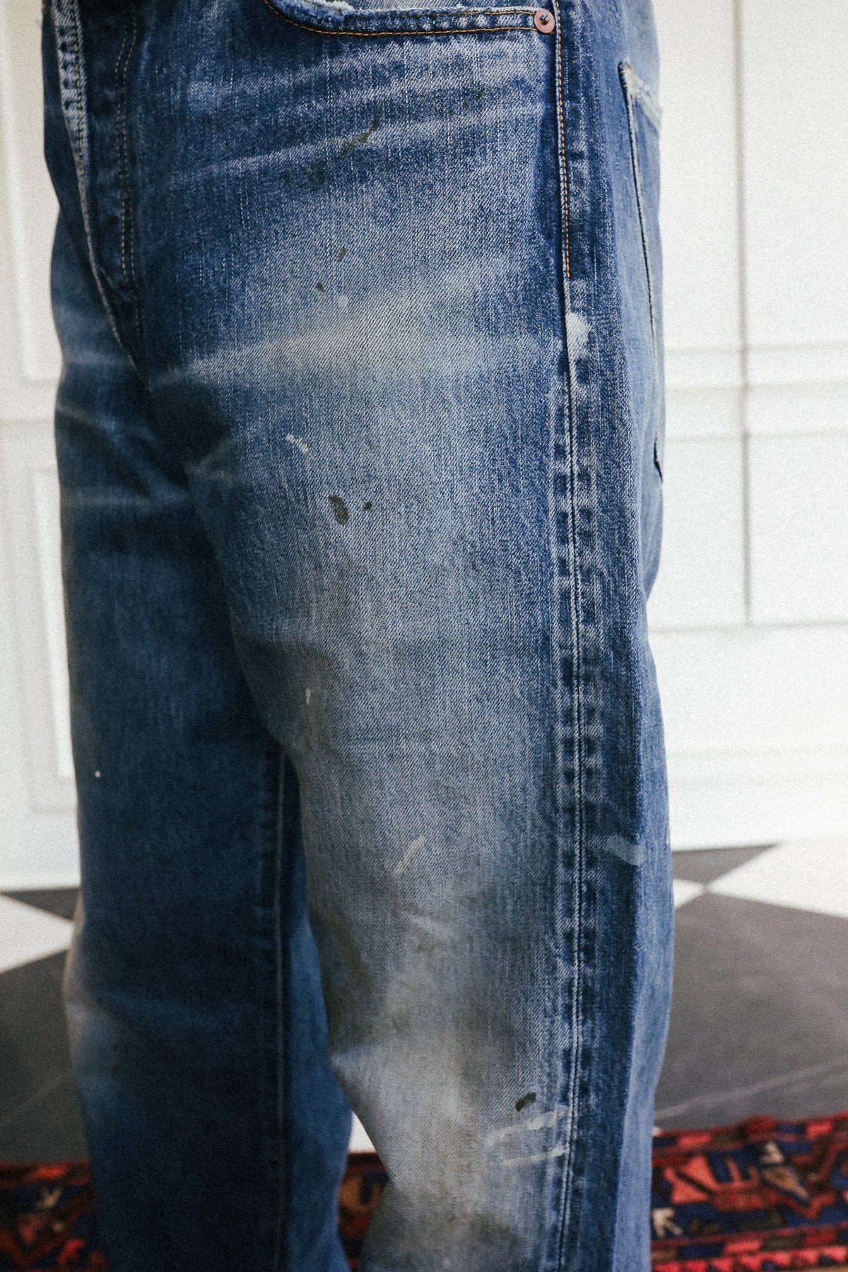 Close-up of a pair of heavily washed, faded blue denim jeans showing significant fading and distressing on the thigh, set against a white paneled wall background.