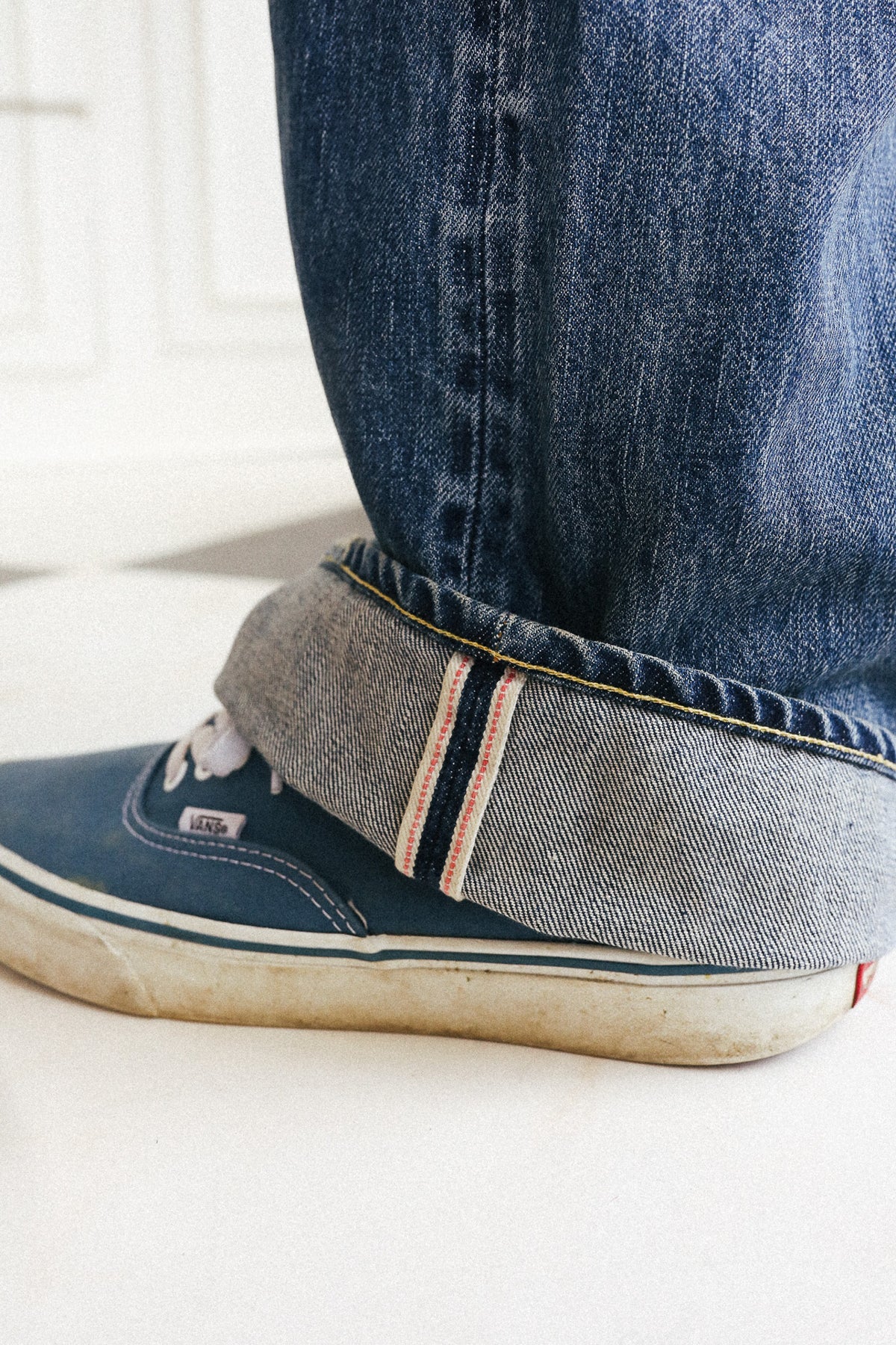 Close-up of a person's foot wearing a navy blue canvas sneaker with the jeans cuff turned up, showing the striped fabric lining and the side tag.