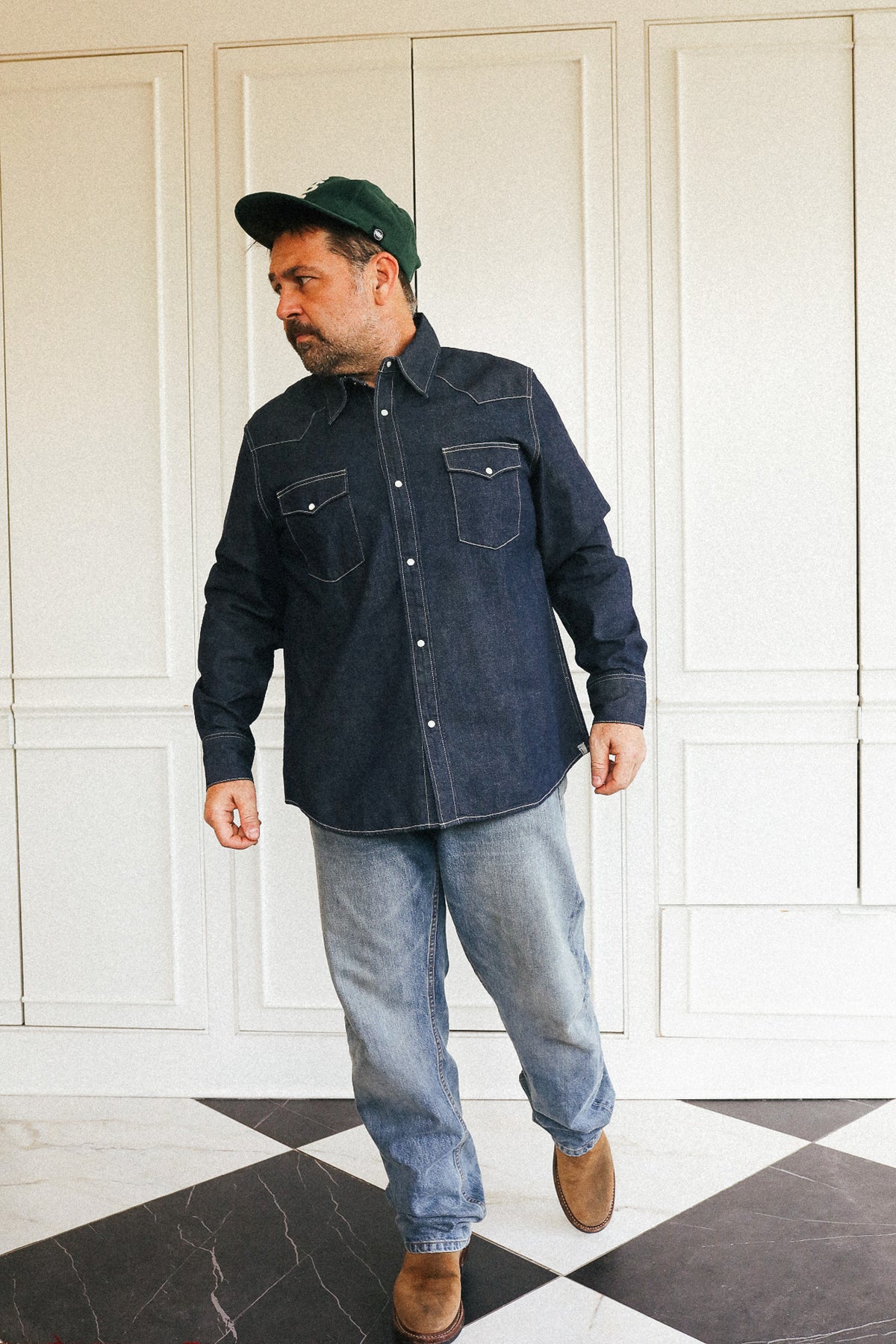 A man wearing a dark indigo denim western-style button-up shirt, light blue jeans, brown boots, and a dark green baseball cap, standing in front of a white paneled wall with a black and white checkered floor.