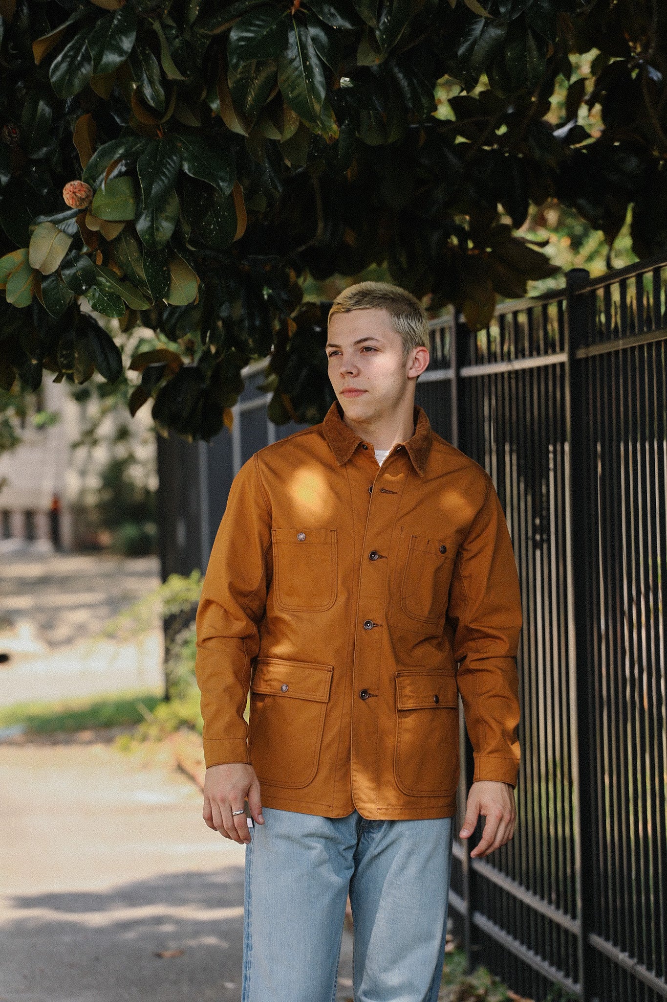 Young man wearing a brown chore jacket and light blue jeans standing by a black metal fence under green foliage
