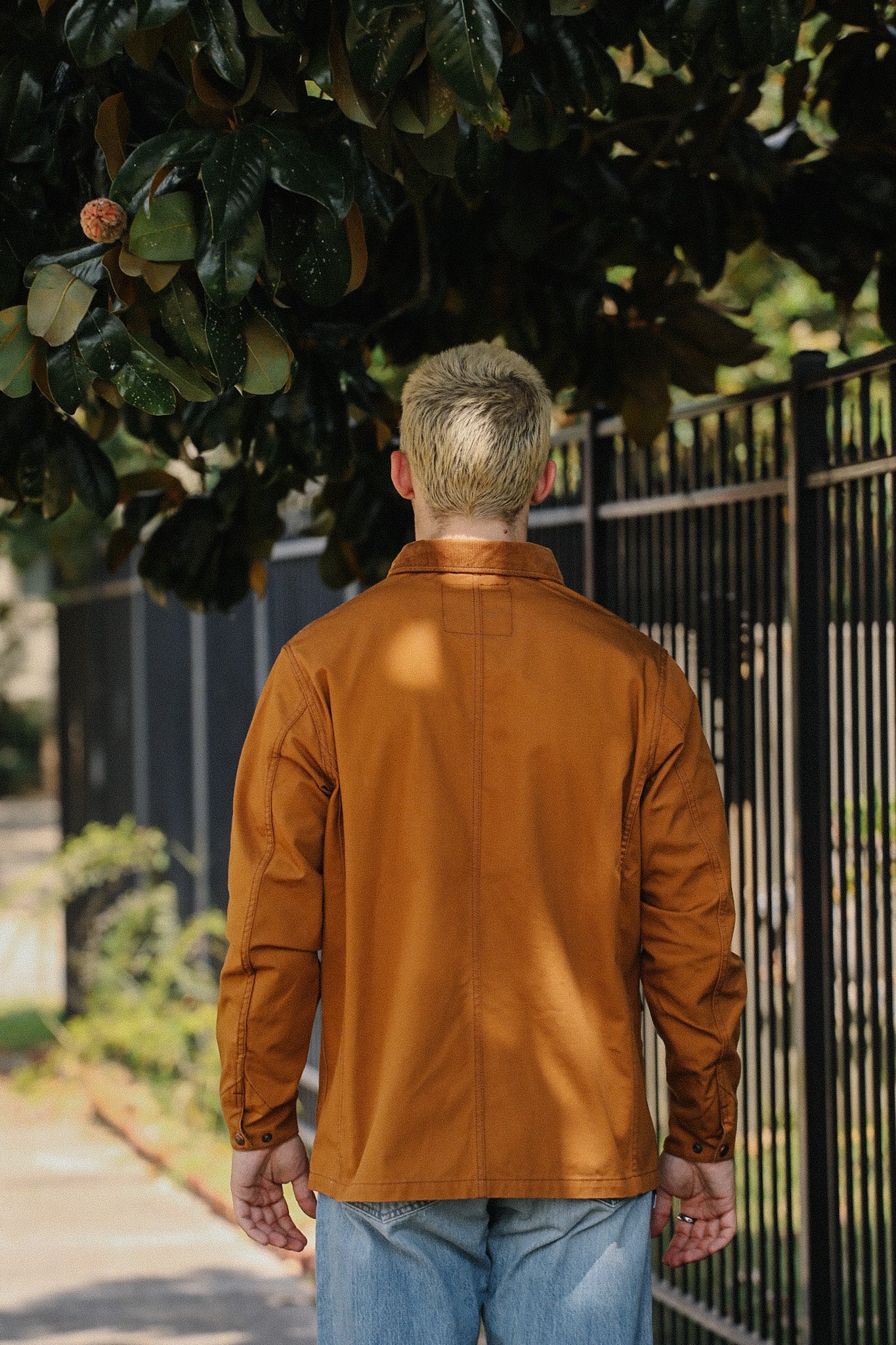 Man wearing a brown Iron Shop Provisions jacket standing outdoors near a black metal fence and large leafy tree