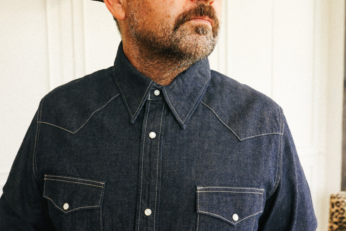 A close-up of a man wearing a dark indigo denim western-style button-down shirt with contrast stitching and two chest pockets.