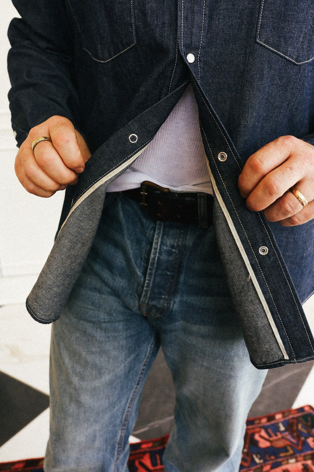 A close-up of a person wearing a dark indigo denim overshirt over a light gray t-shirt and blue jeans, with the overshirt being opened to show the inner lining.