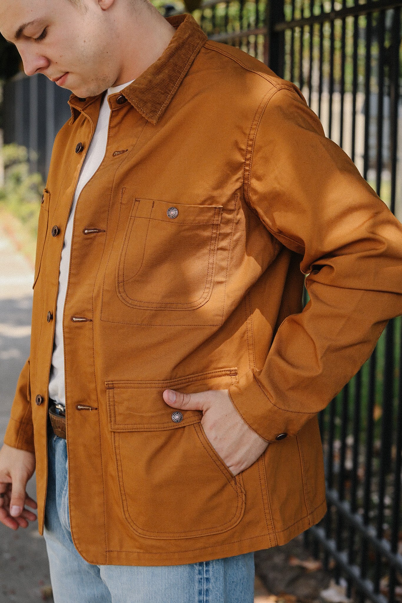 Man wearing brown workwear jacket with large pockets and corduroy collar outdoors by metal fence