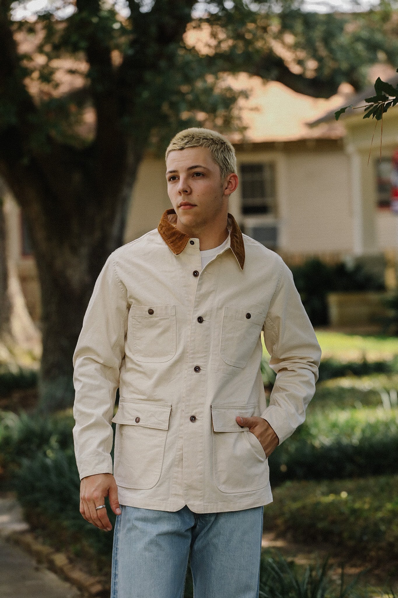 Young man wearing cream chore jacket with brown collar and light blue jeans outdoors