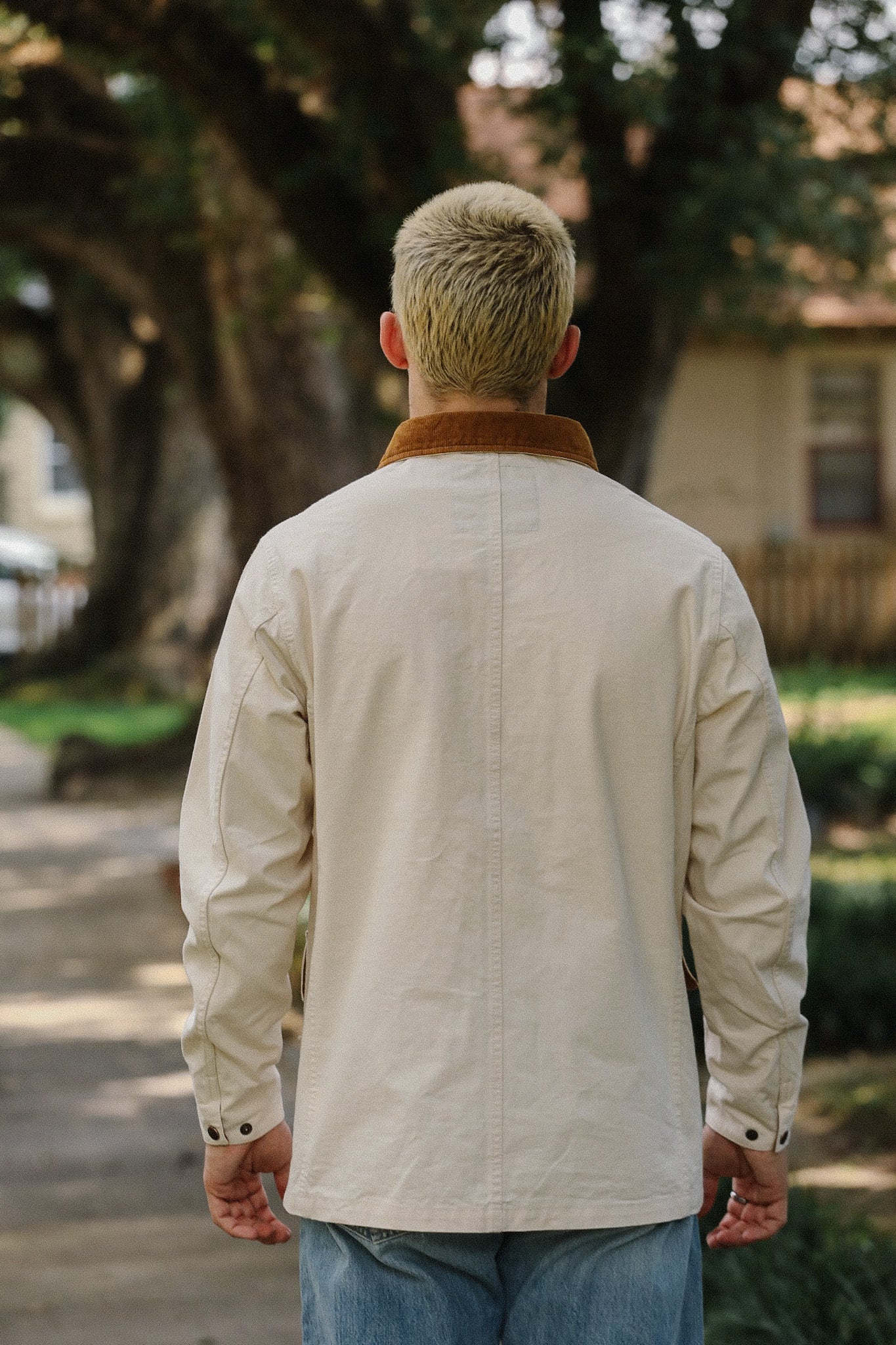 Back view of man wearing cream canvas chore jacket with brown collar and blue jeans outdoors