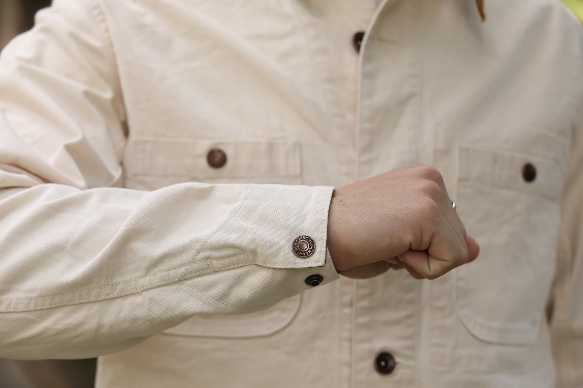 Close-up of a beige work jacket sleeve and cuff with brown metal snap button, worn by a person with a clenched fist