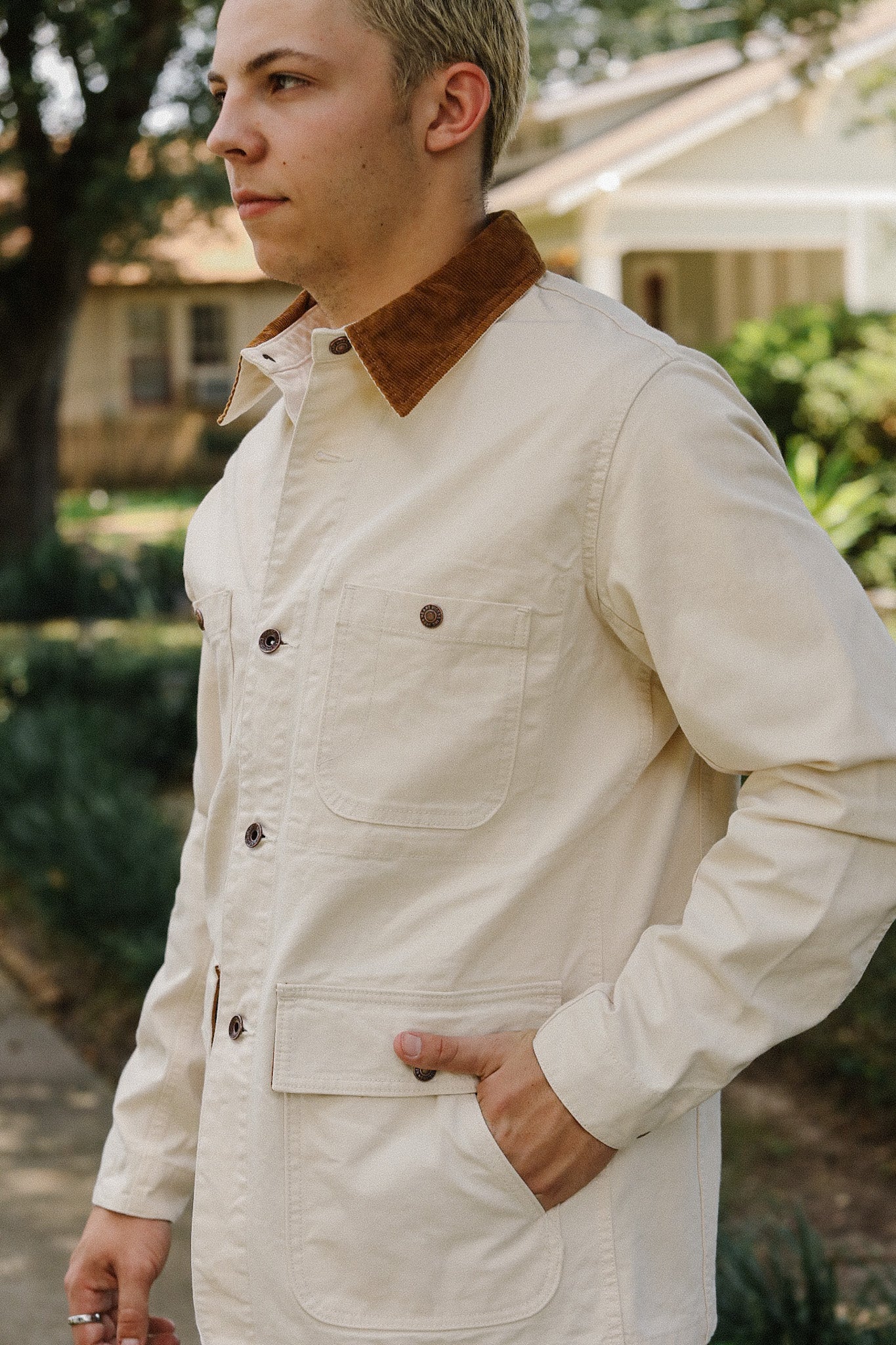 Man wearing a cream work jacket with brown corduroy collar, buttoned pockets, standing outdoors near house
