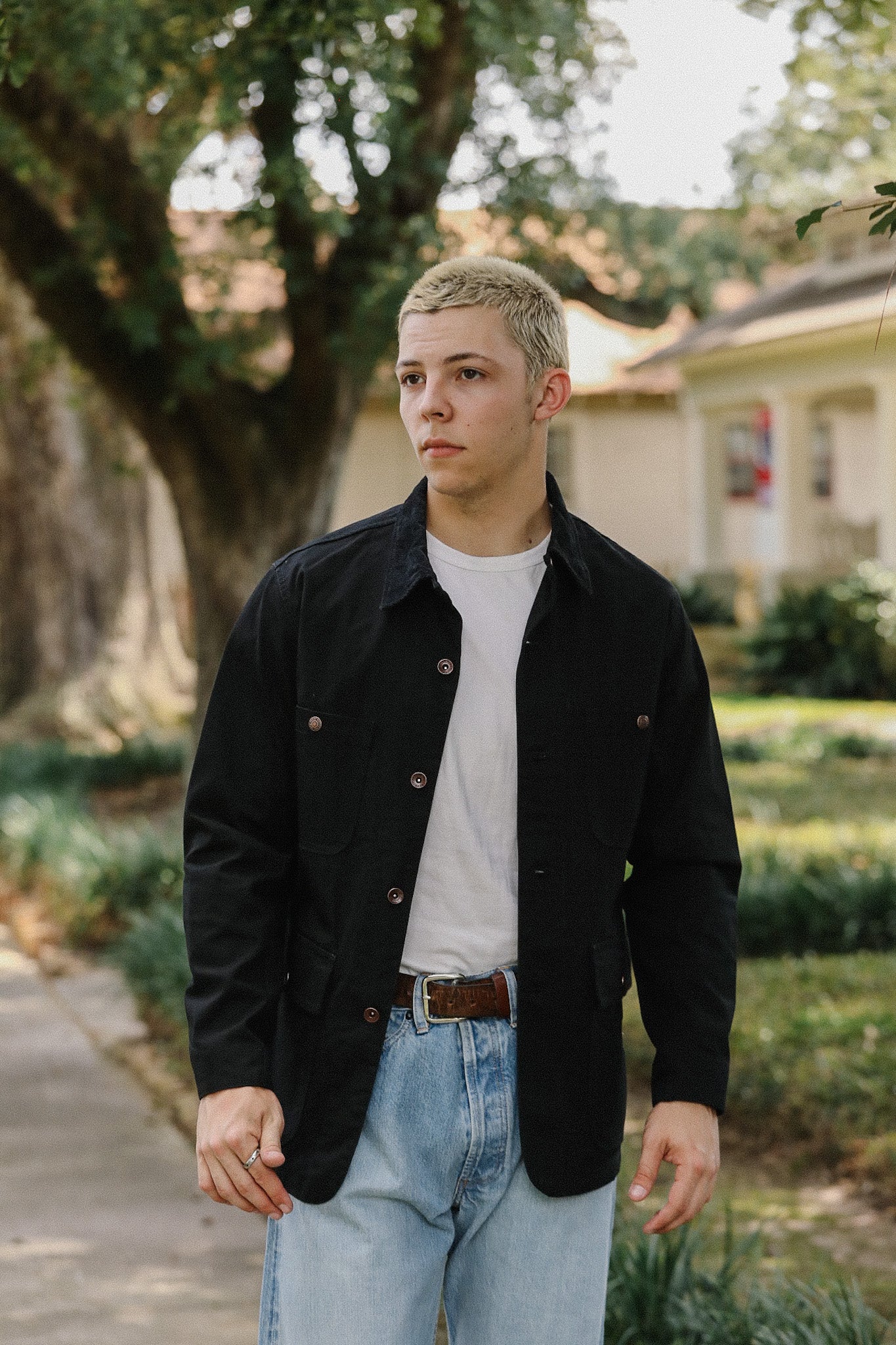 Young man with blonde hair wearing a black button-up jacket, white t-shirt, and light blue jeans outdoors