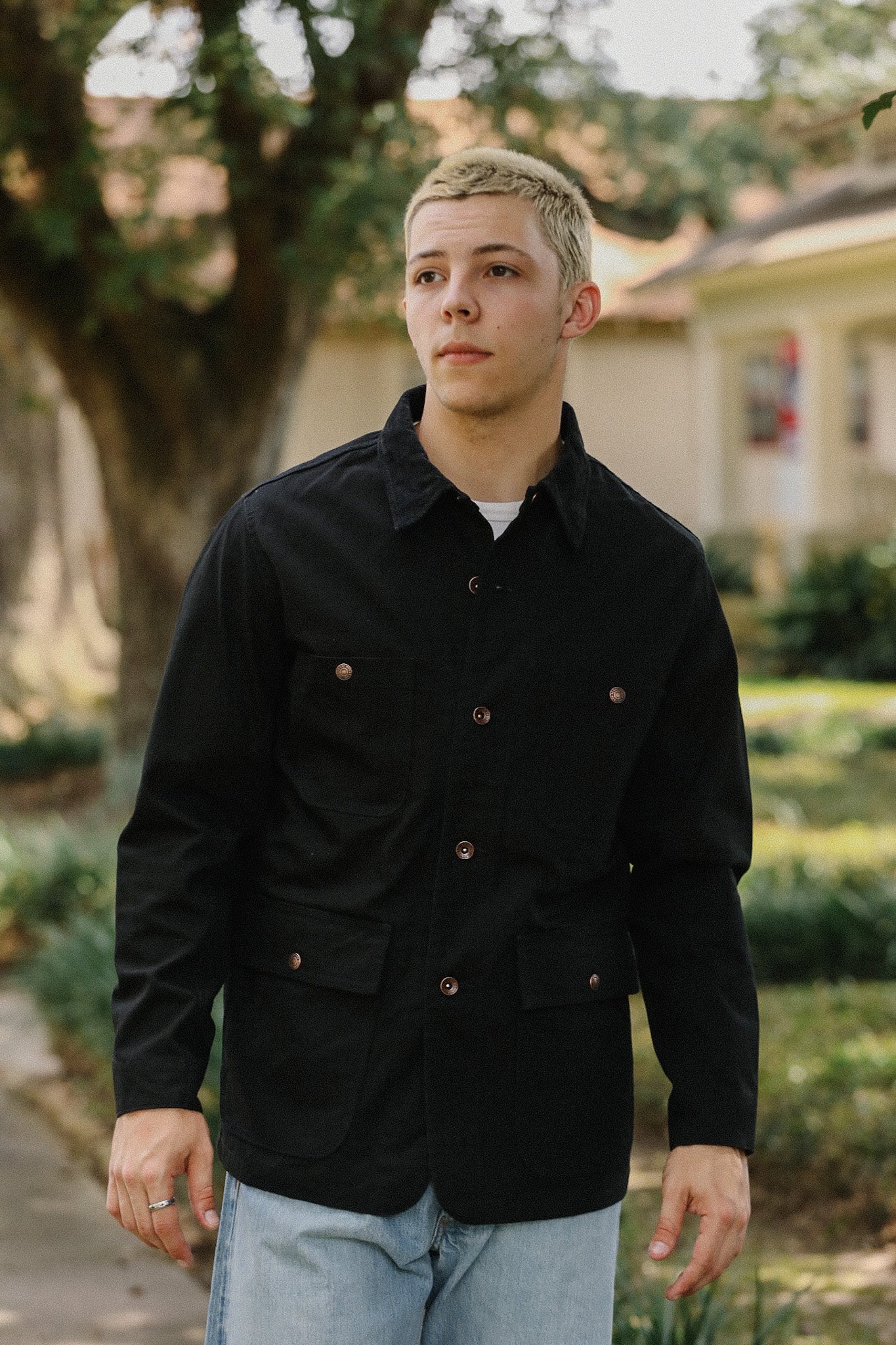 Young man wearing a black button-up jacket with pockets and light blue jeans outdoors near trees