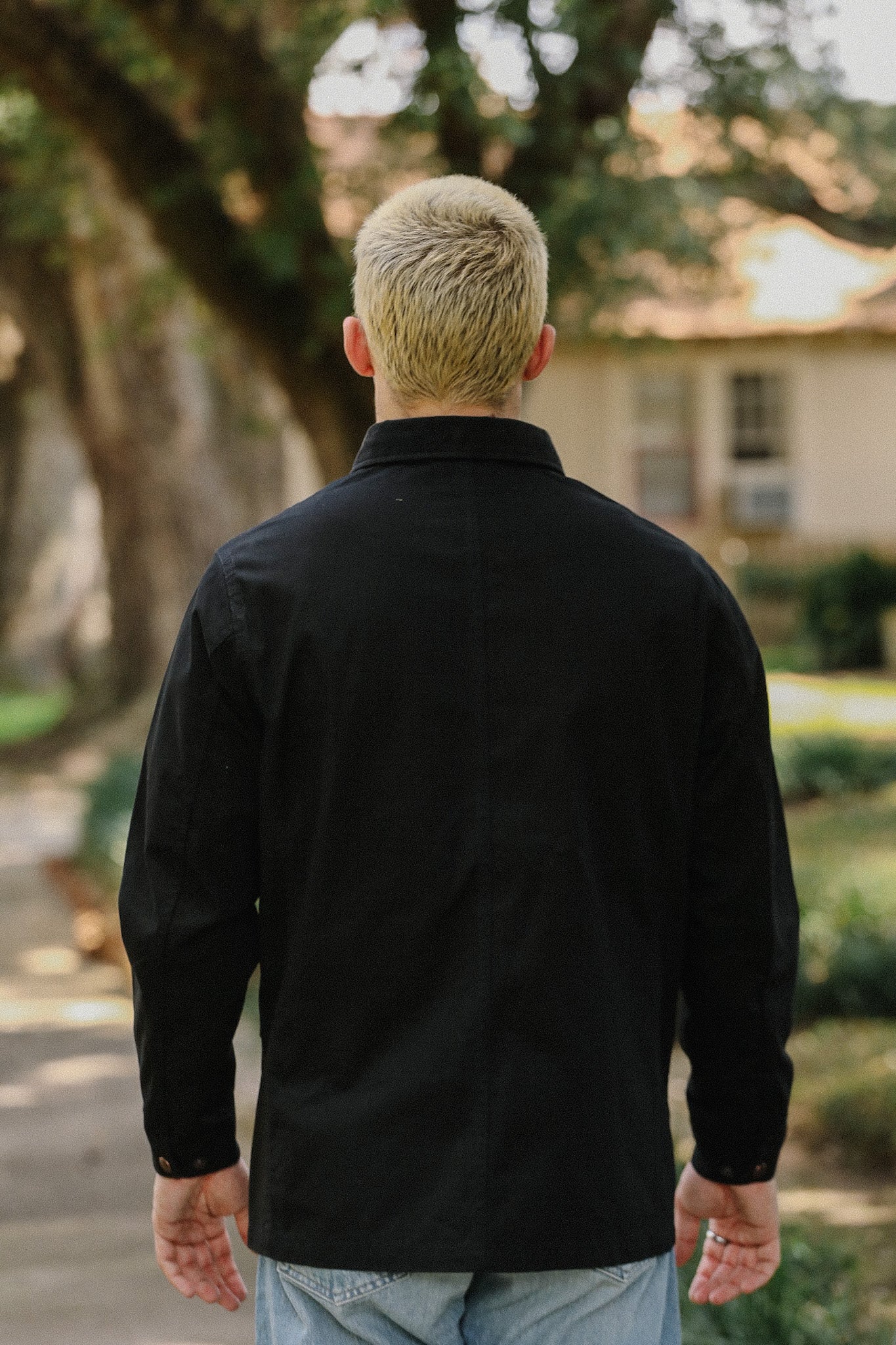 Man with blonde hair wearing a black jacket and blue jeans standing outdoors with trees and a house in background