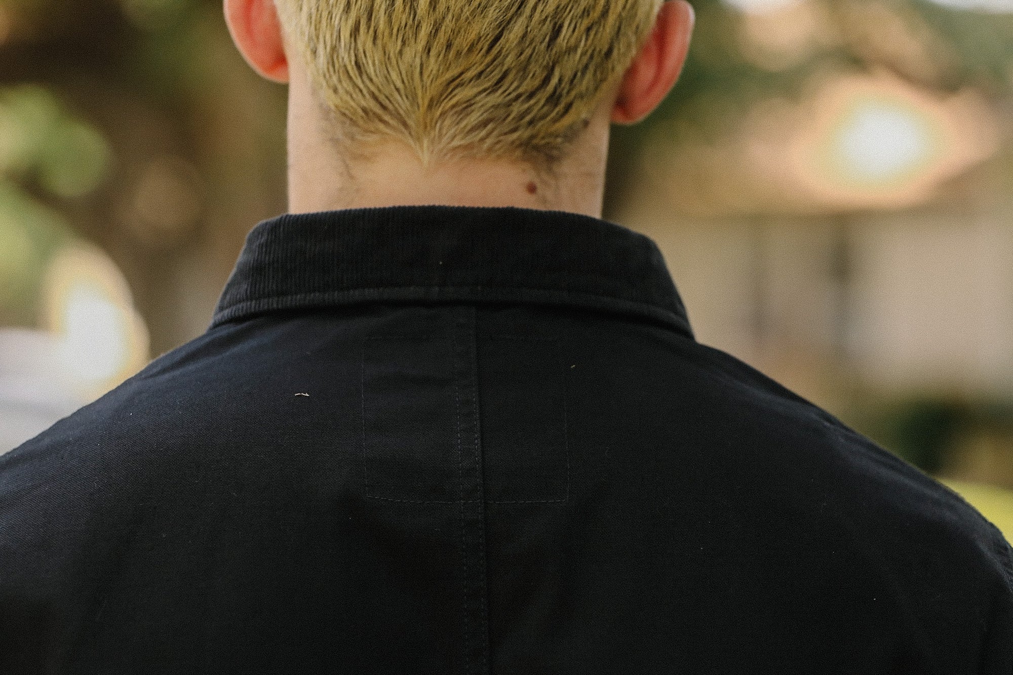 Back view of person with short blonde hair wearing a black collared jacket outdoors