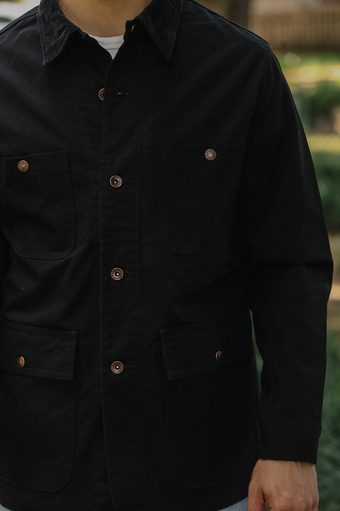 Close-up of a person wearing a black work jacket with four pockets and copper buttons, outdoors