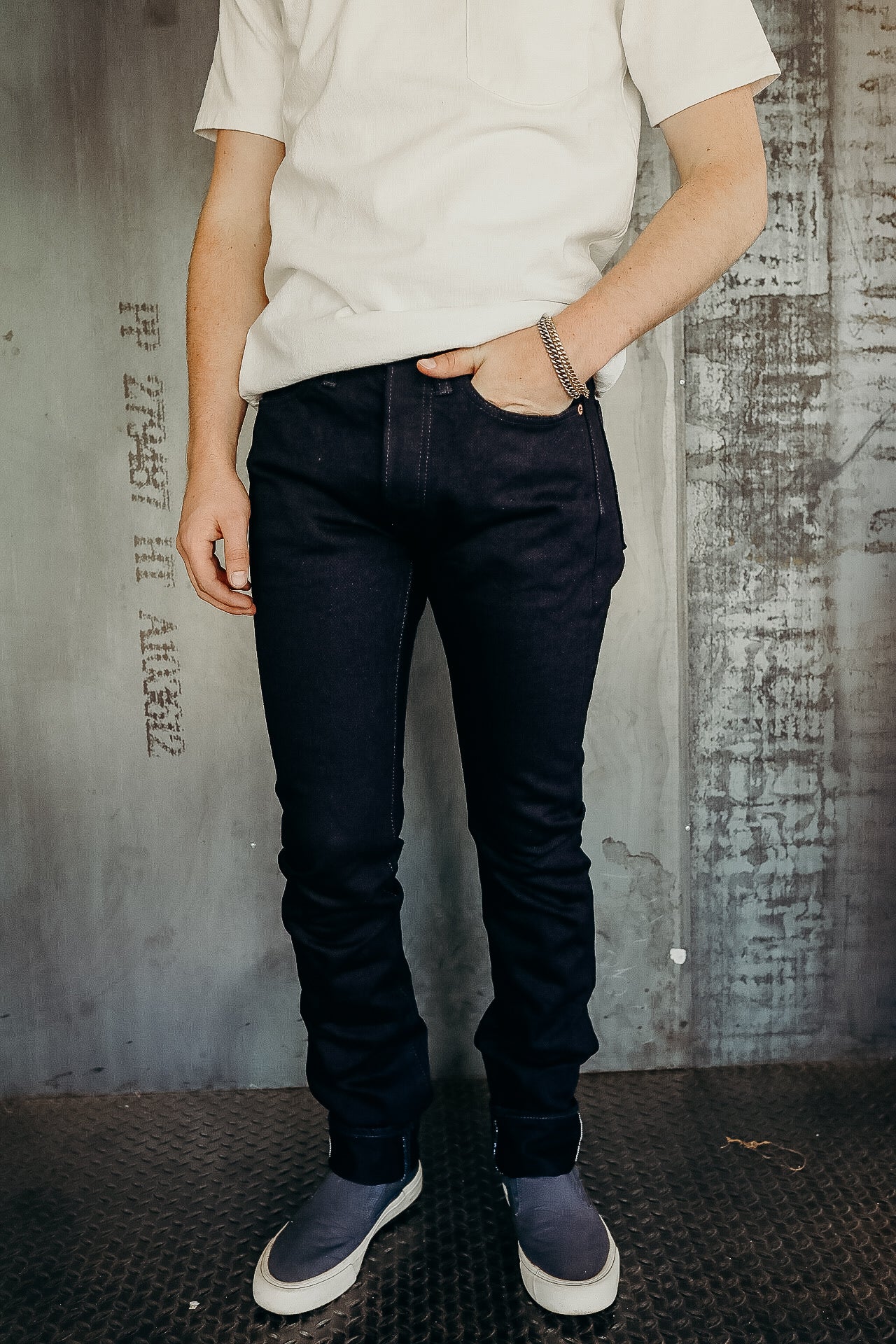 Man wearing dark raw denim jeans, white short-sleeve shirt, and gray slip-on shoes, standing on industrial floor
