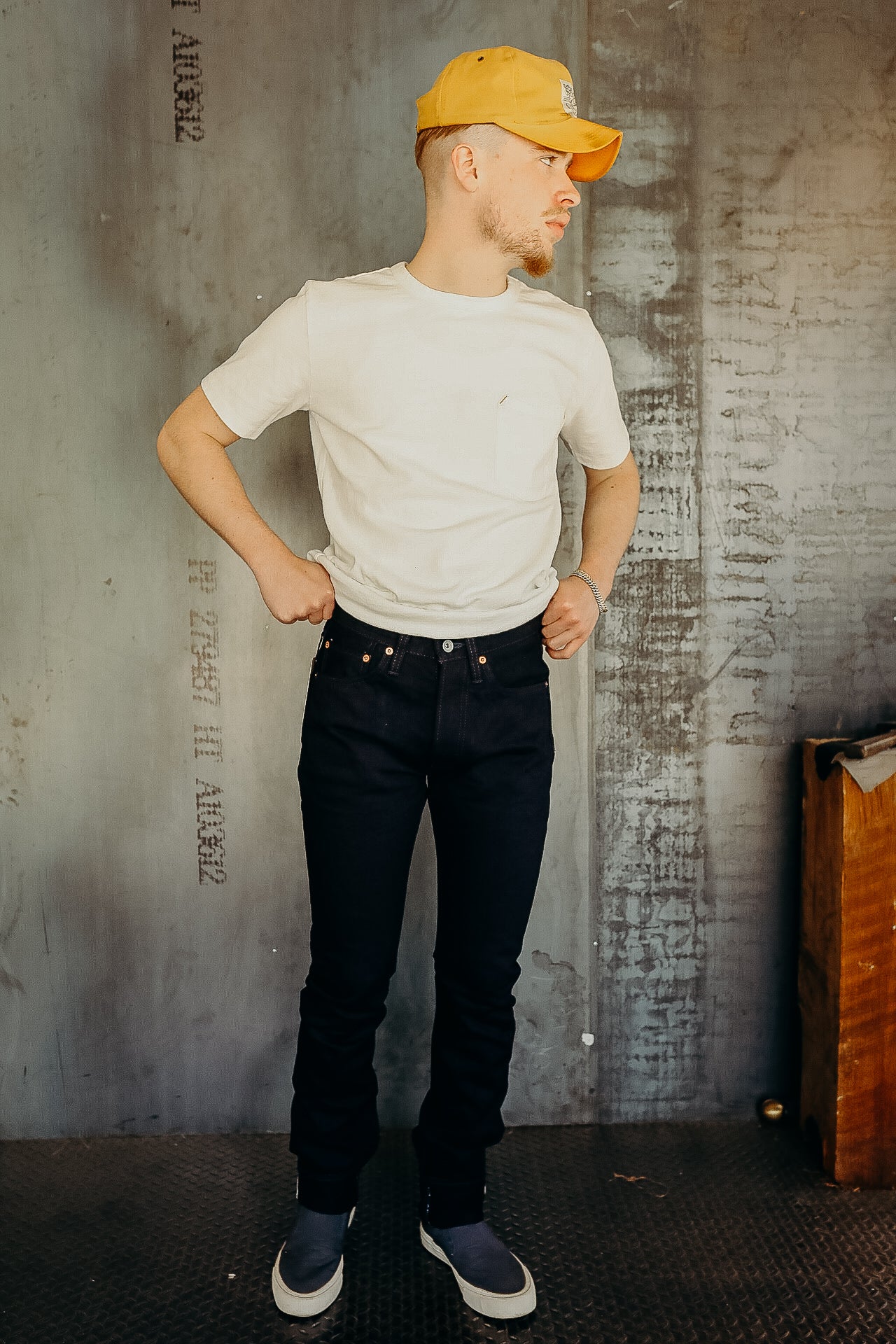 Man wearing yellow cap, white t-shirt, black jeans, and slip-on shoes in industrial setting