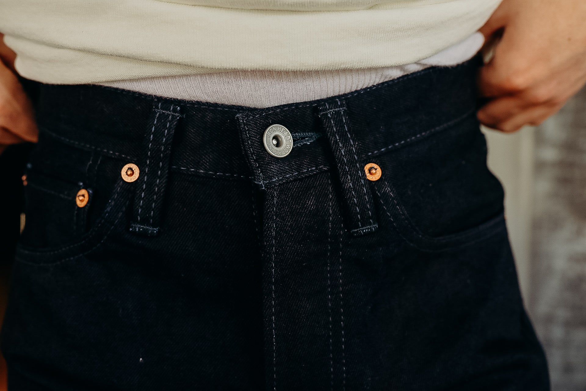 Close-up of black denim jeans waist with visible button and copper rivets, styled with tucked beige shirt
