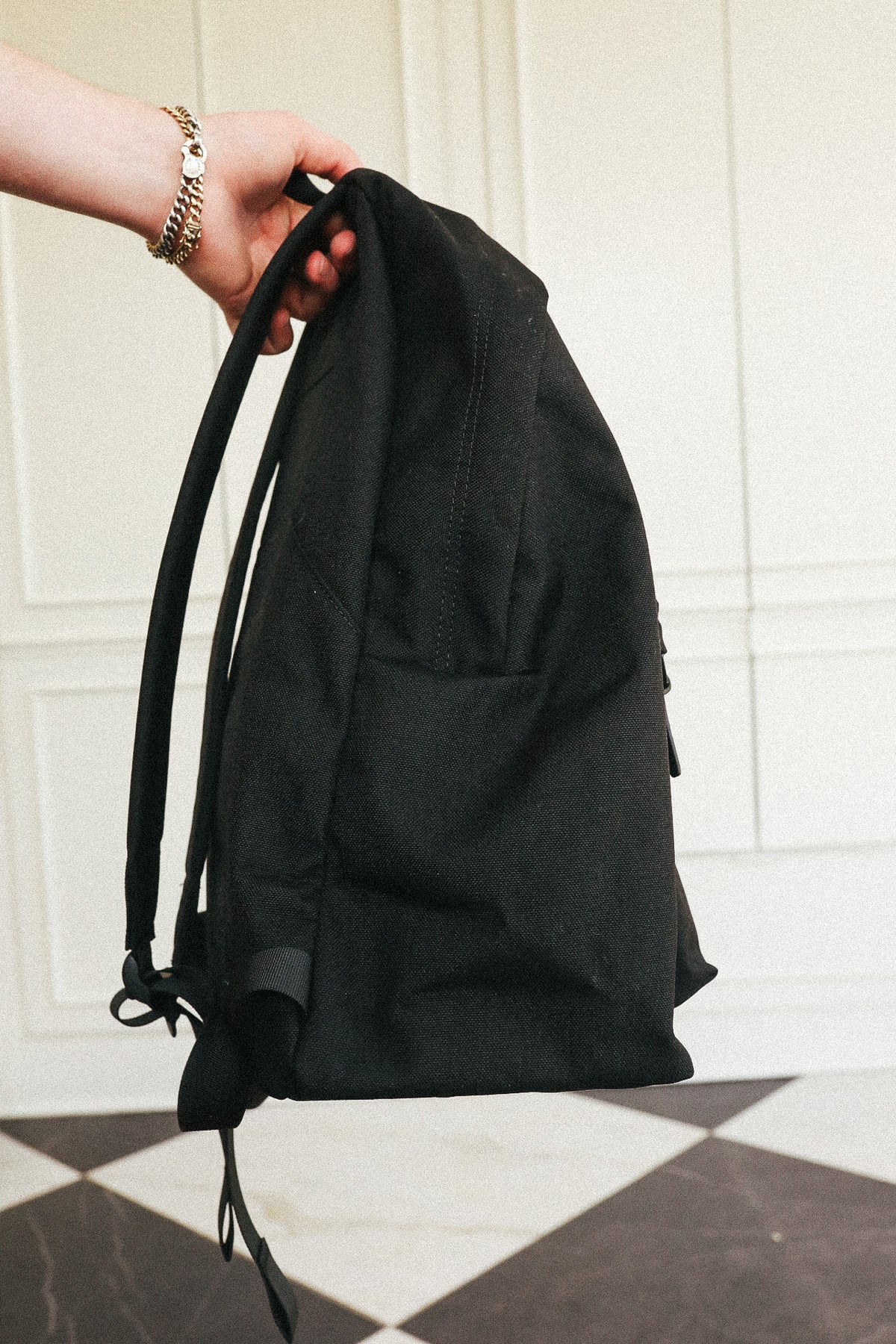 A black canvas drawstring backpack held by a hand wearing a gold chain bracelet, set against a white paneled wall and a black-and-white checkered floor.