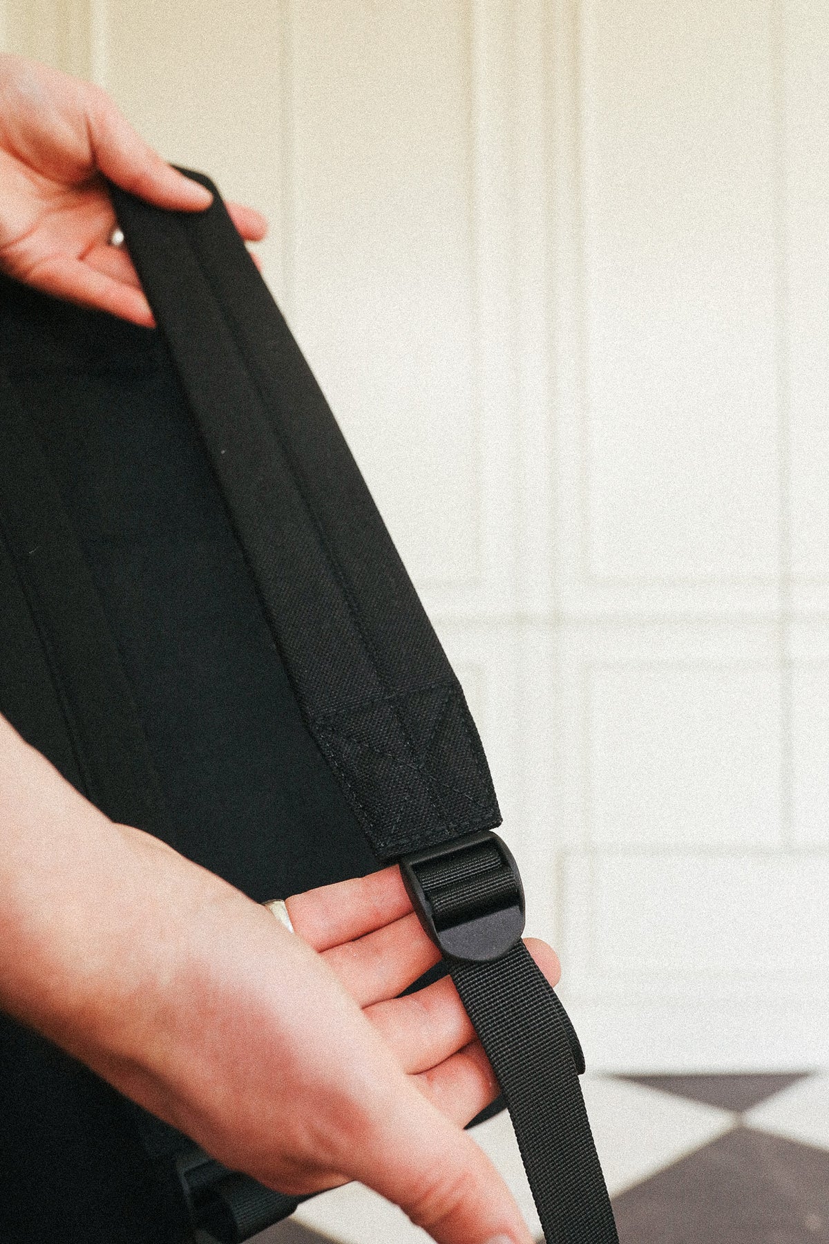 Close-up of a person's hands adjusting a black adjustable shoulder strap with a plastic quick-release buckle on a black bag, set against a light paneled wall background.