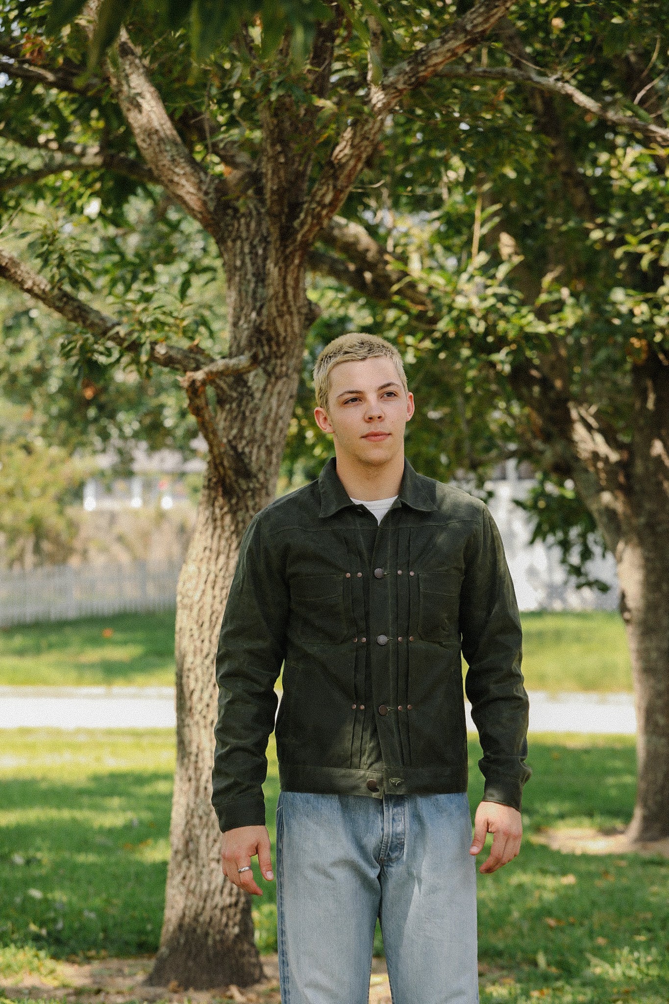 Young man wearing dark green buttoned jacket and light blue jeans standing outdoors near leafy trees