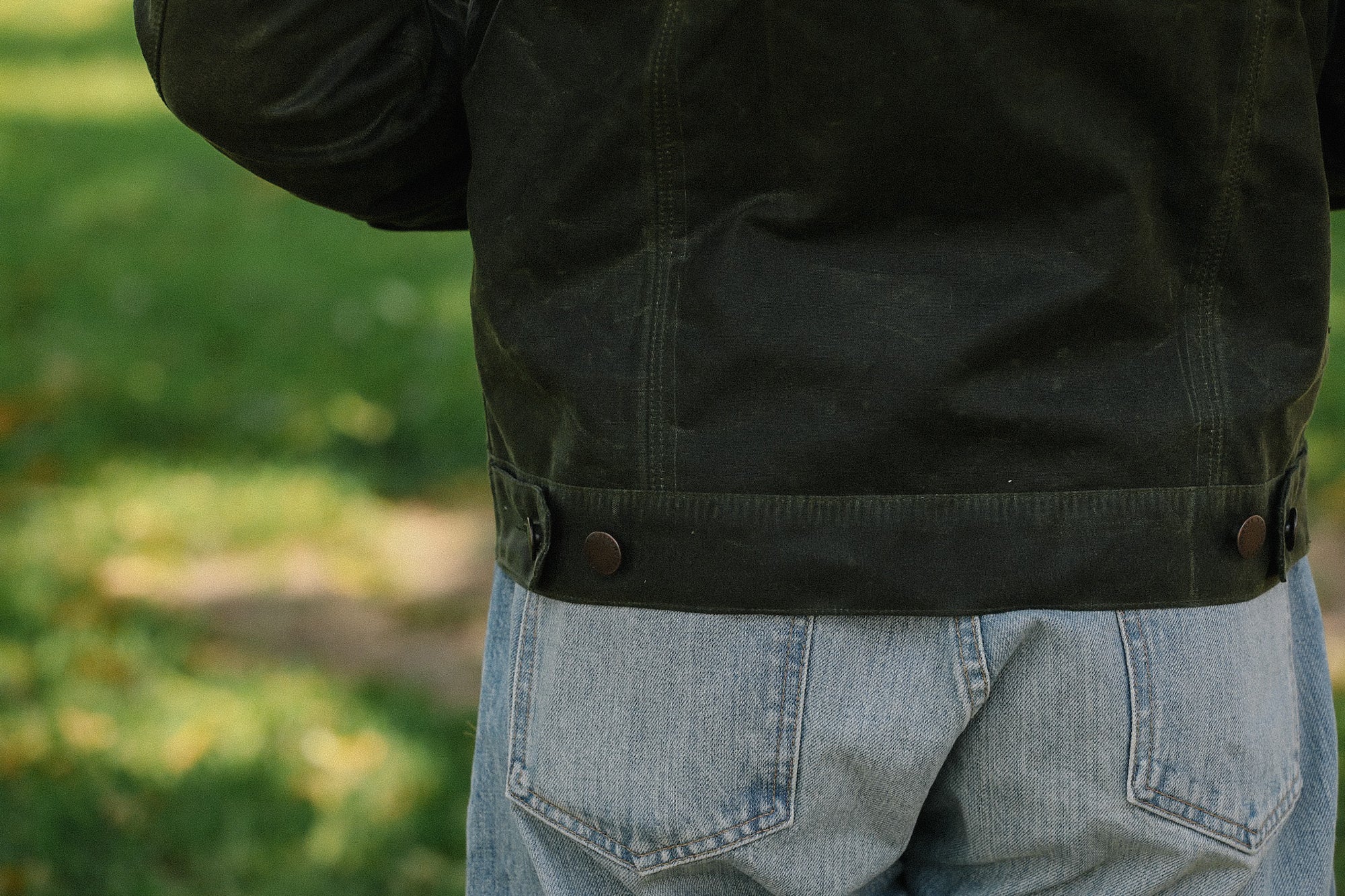 Back view of person wearing black waxed canvas jacket and light blue denim jeans outdoors