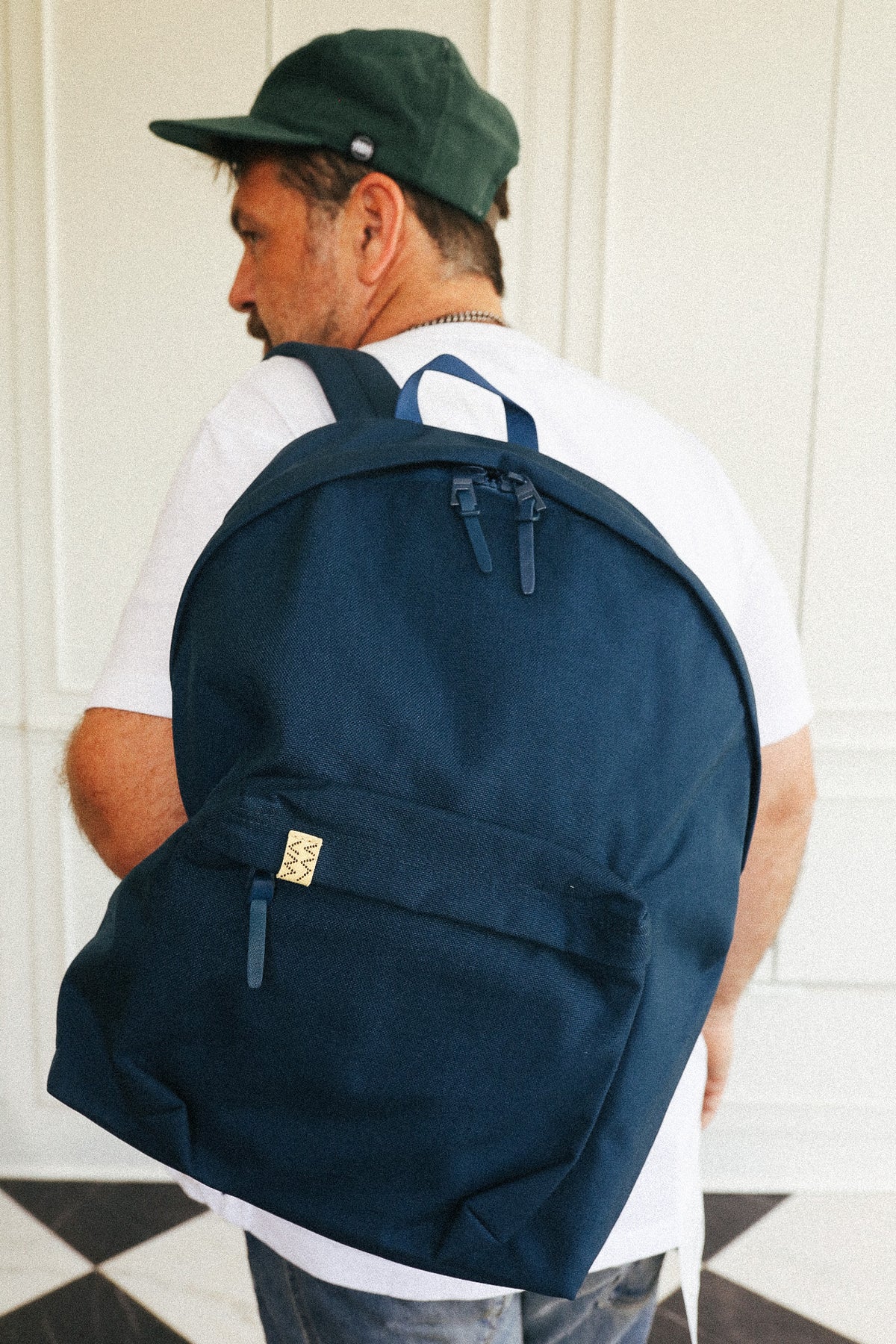 A man wearing a dark navy blue canvas backpack with a front zippered pocket and a small gold brand tag, photographed from behind against a white paneled wall.