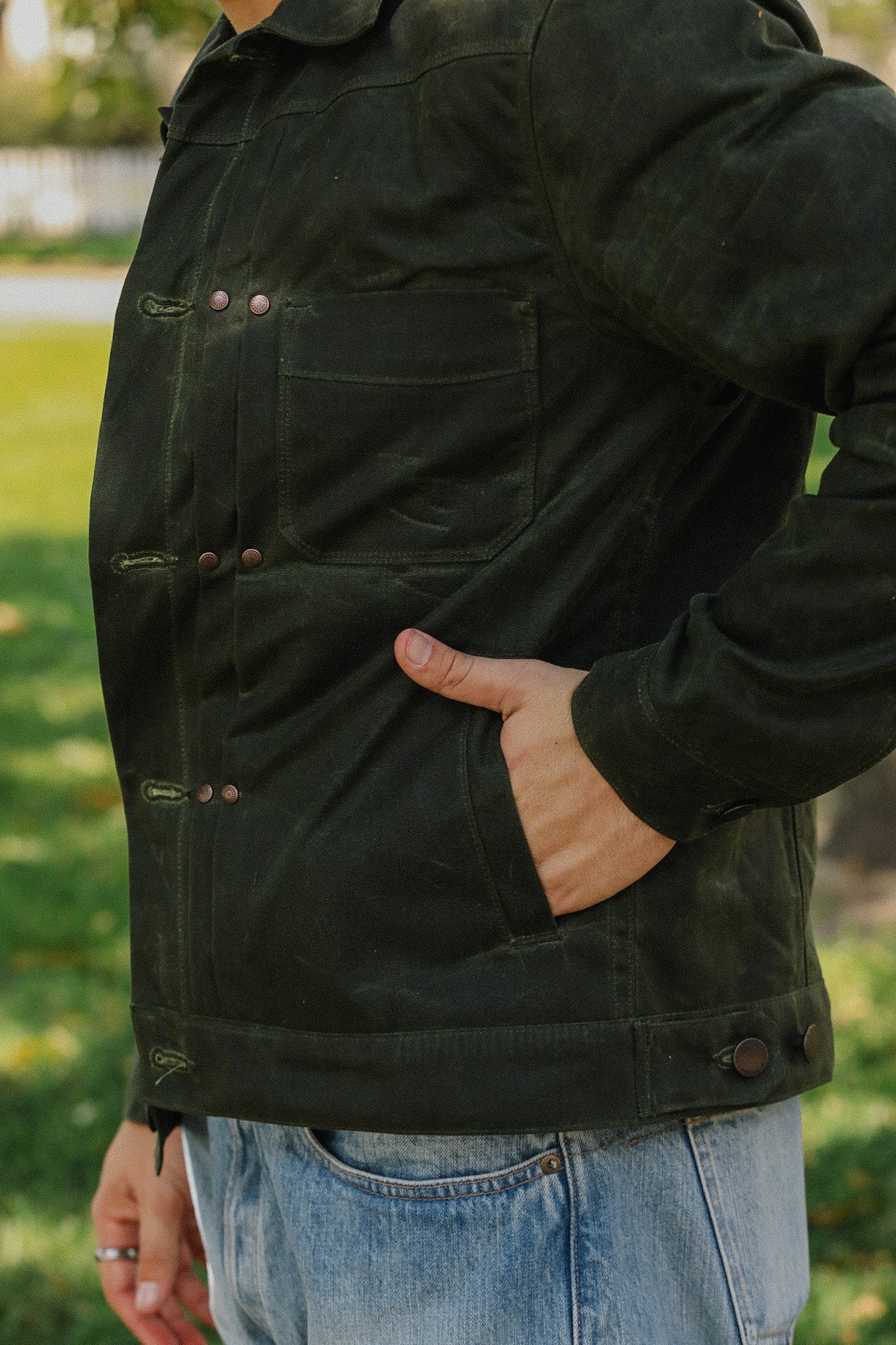 Close-up of a person wearing a dark green waxed canvas jacket with copper buttons and light blue jeans outdoors