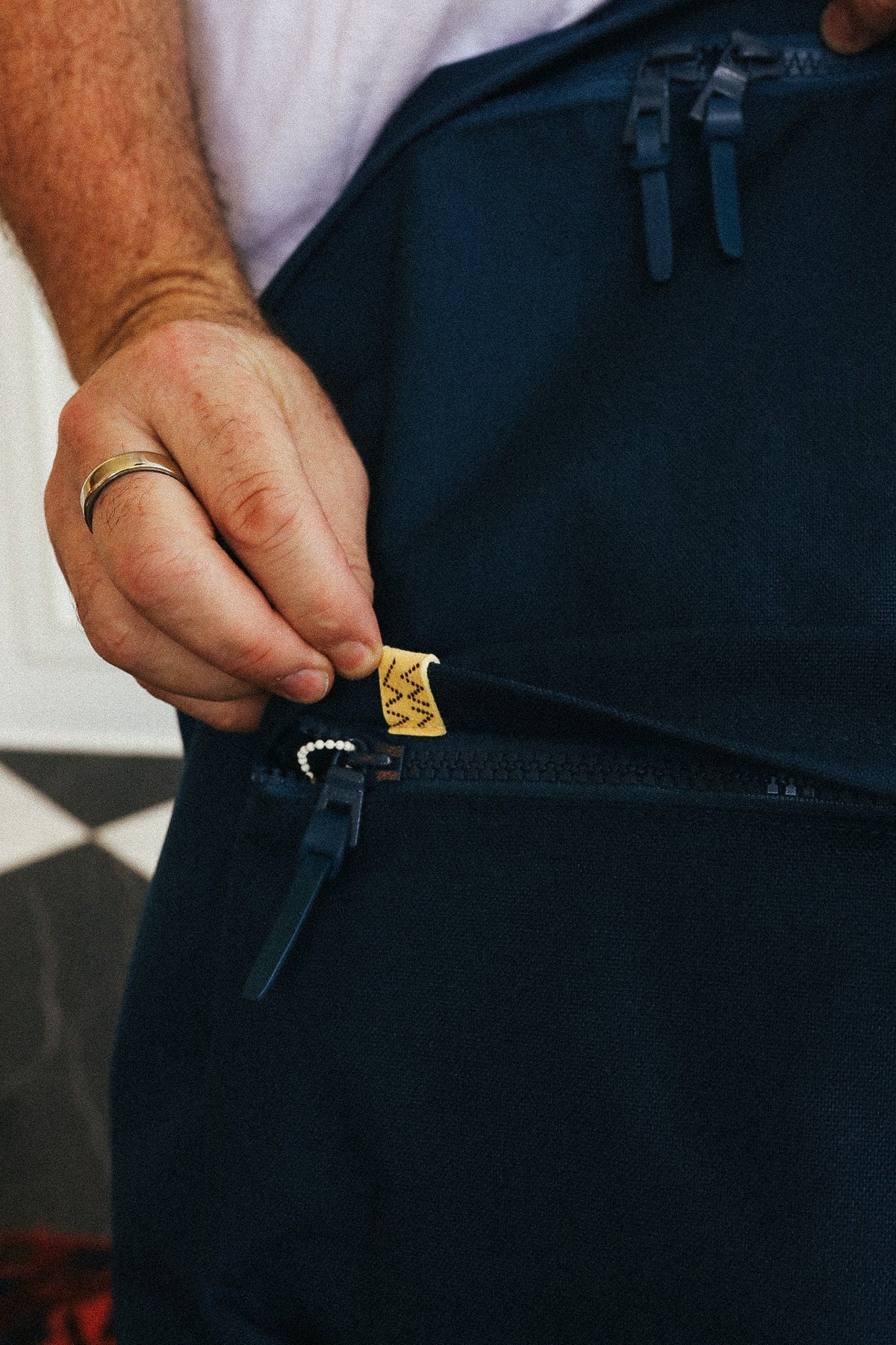 Close-up of a person's hand opening a zipper on a dark navy blue apron, with a small gold brand tag visible.
