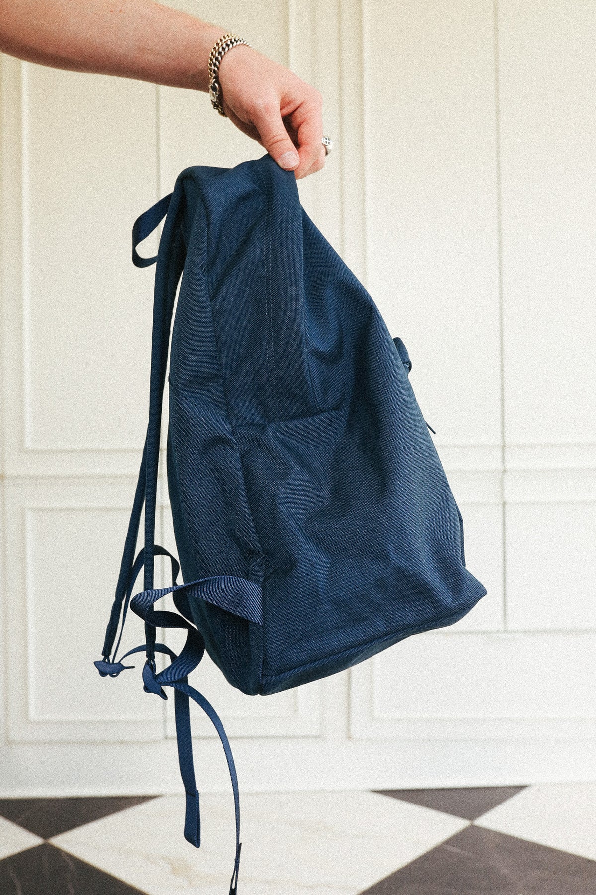 A navy blue canvas backpack held by a person's hand in front of a white paneled wall and black-and-white checkered floor.