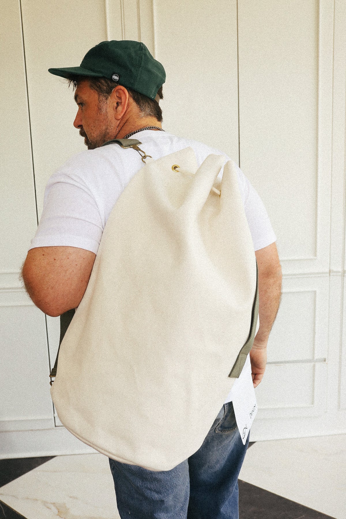 A man wearing a white t-shirt, blue jeans, and a dark green baseball cap is shown from behind, carrying a large, natural canvas drawstring backpack on his shoulder in front of a white paneled wall.