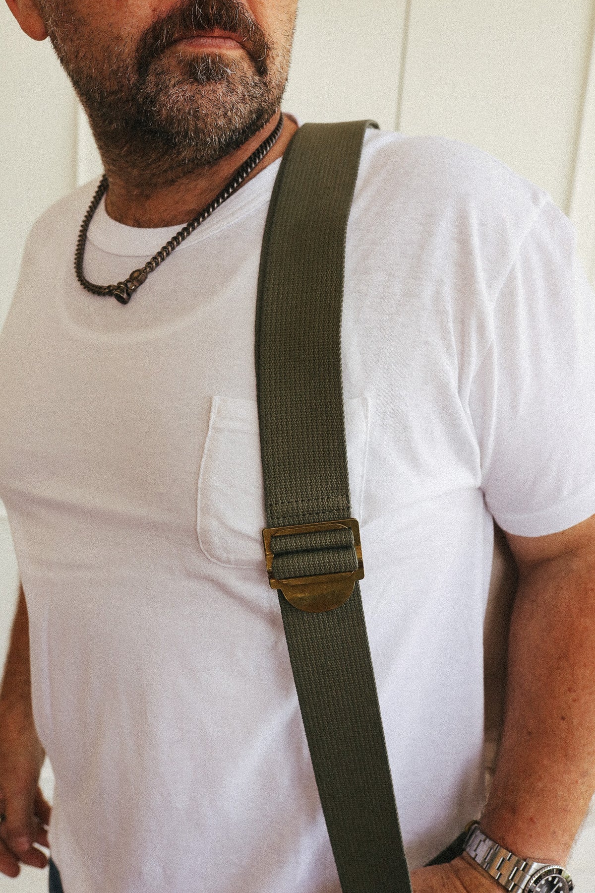 A close-up of a man wearing a white t-shirt and a dark olive green canvas shoulder strap with a bronze-toned metal buckle, likely for a bag or crossbody accessory.