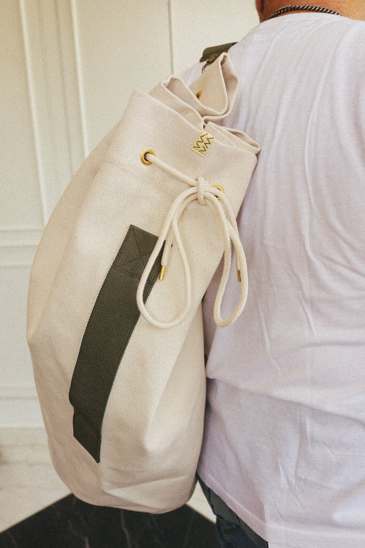 A person wearing a white t-shirt carries a large cream canvas backpack with an olive green vertical stripe and gold-tone hardware.