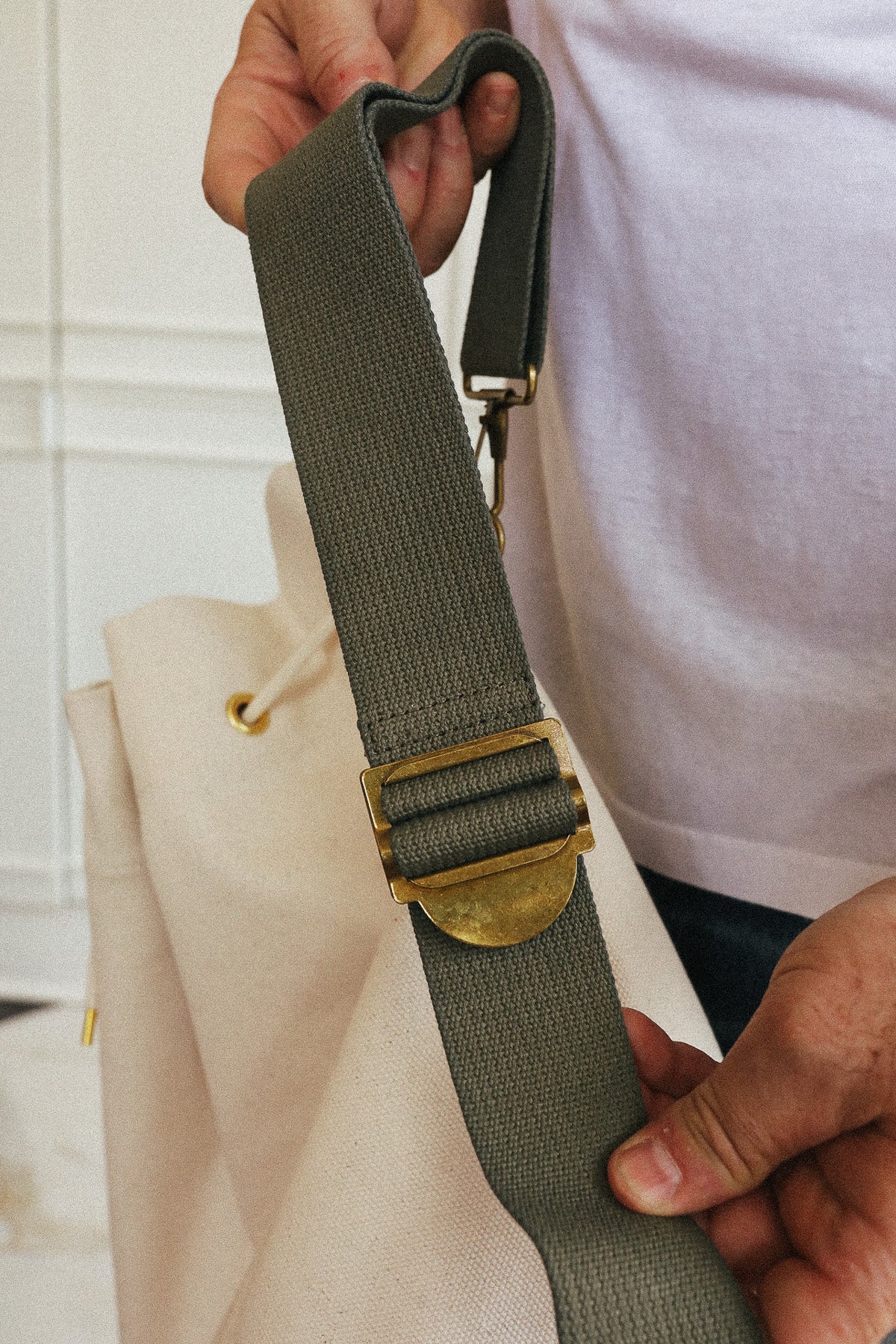 Close-up of a person holding a beige canvas tote bag with an olive green adjustable shoulder strap and a brass buckle.