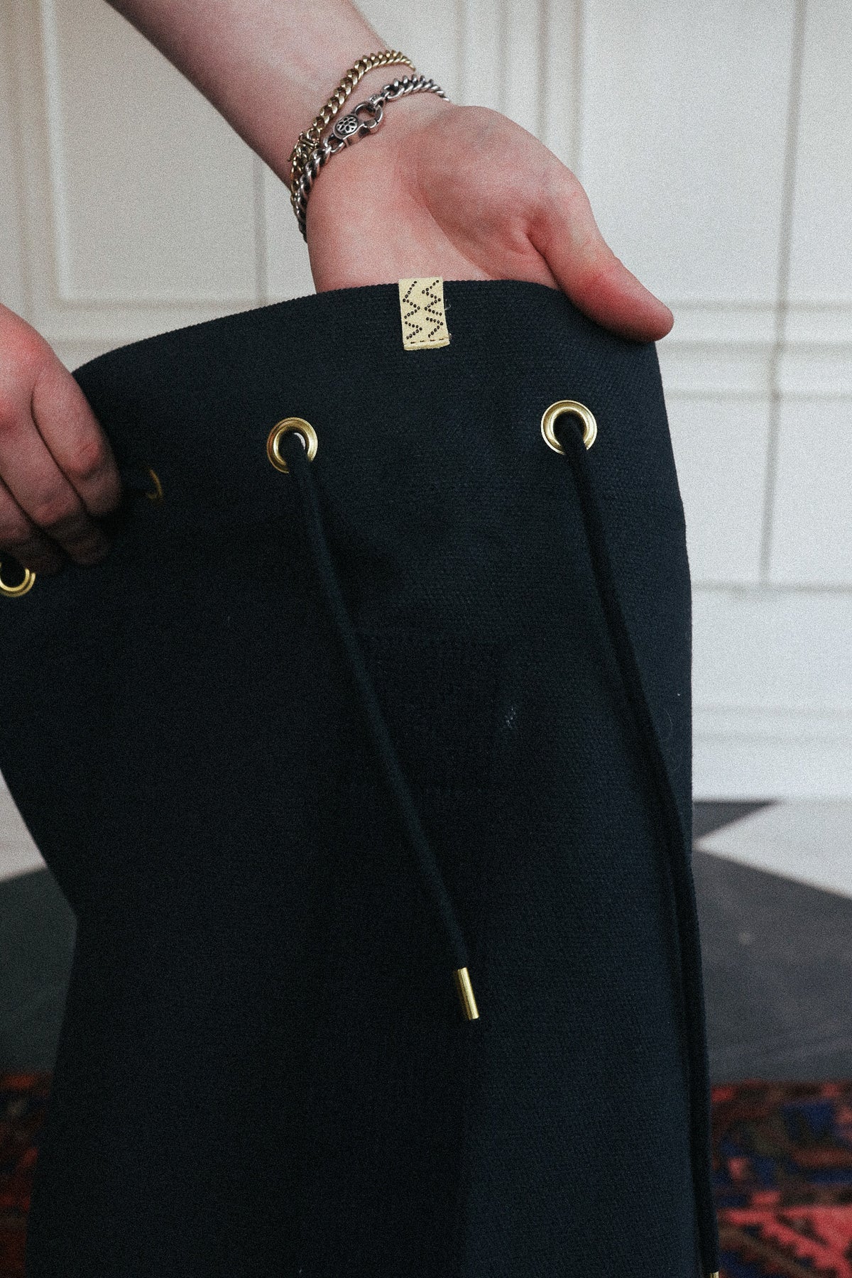 A close-up of a person holding a black drawstring bag with gold-tone metal grommets and a small gold geometric logo tab near the top.