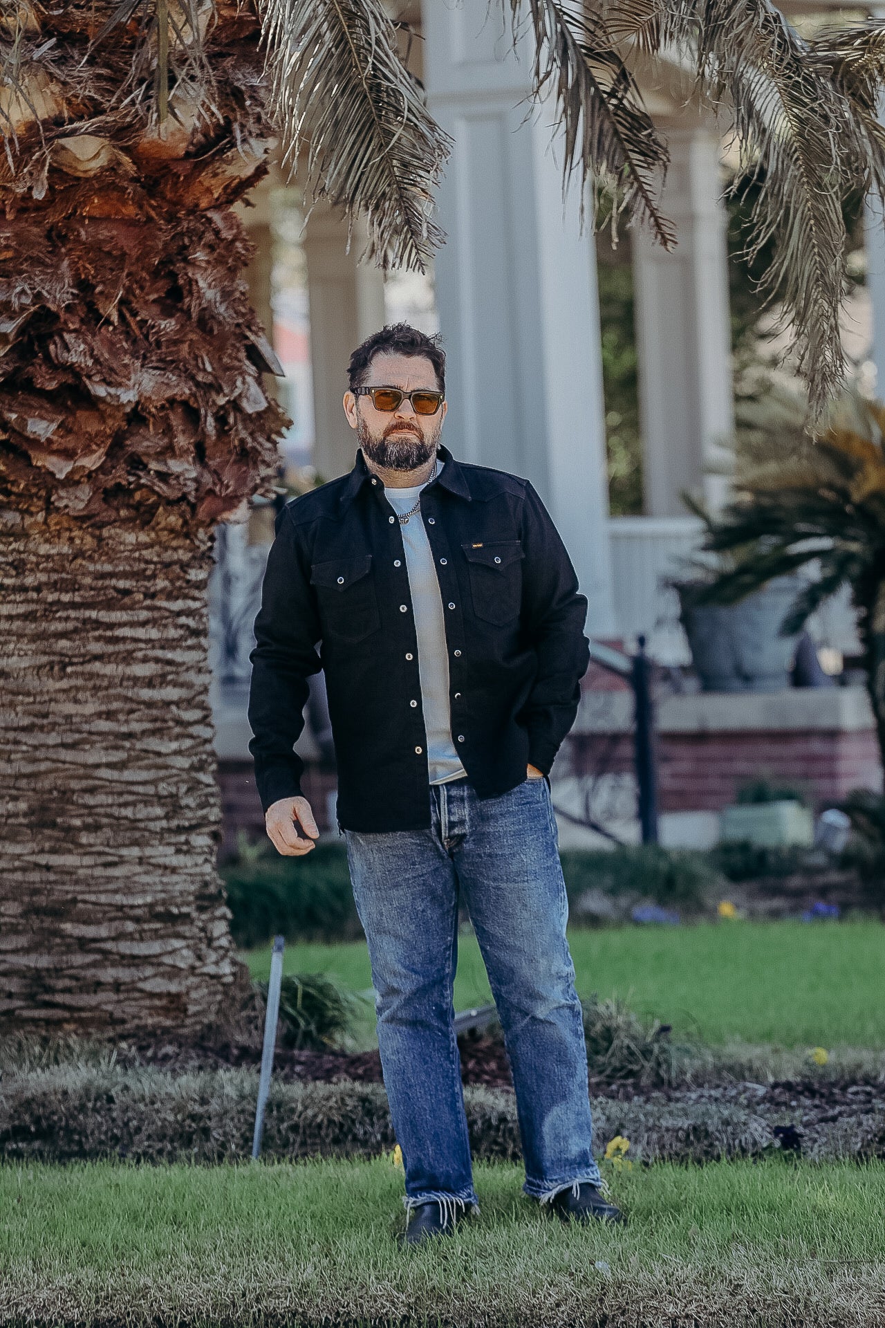 Man with beard and sunglasses wearing black jacket and blue jeans standing on grass near palm tree