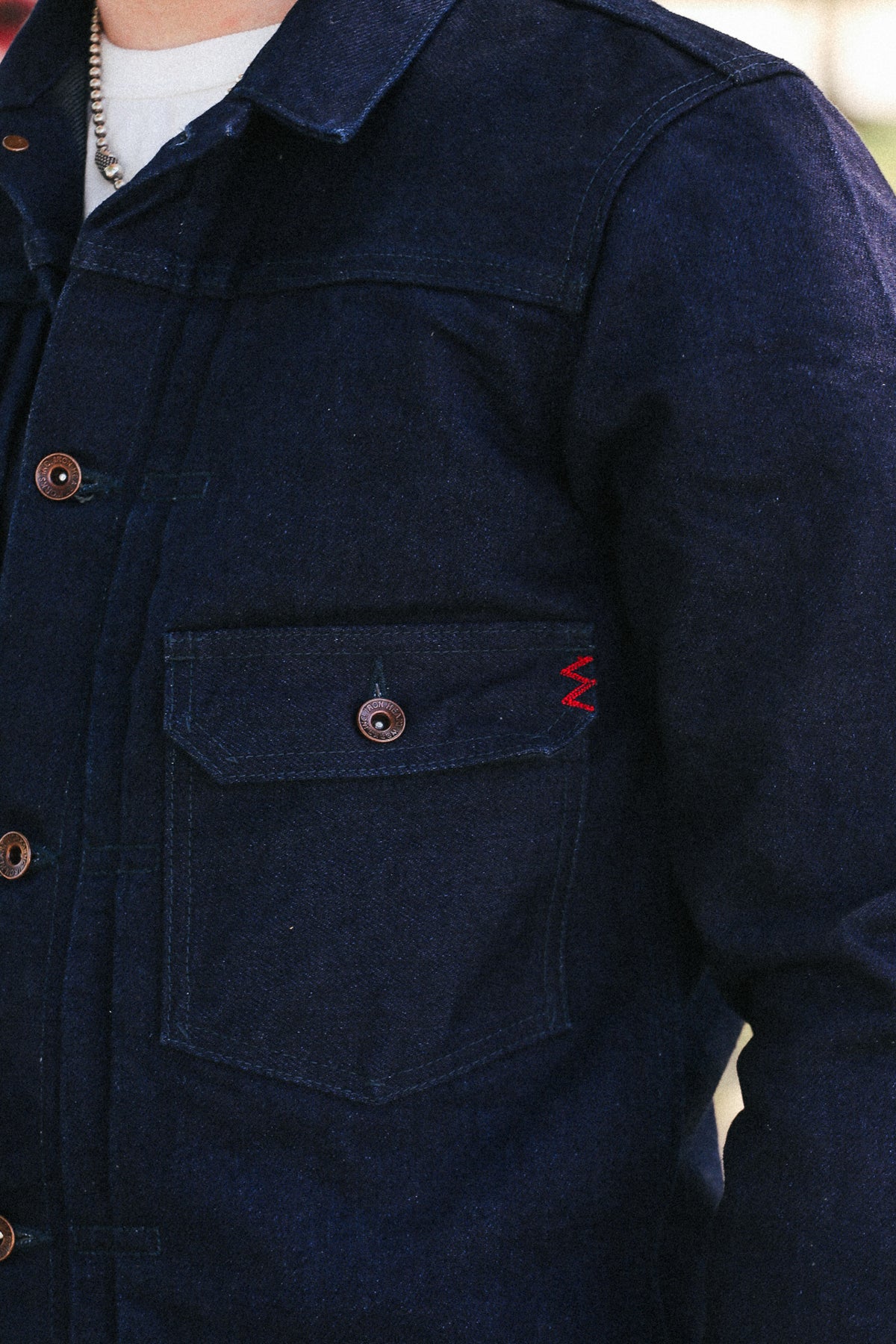 Close-up of a person wearing a dark indigo denim jacket with a chest pocket, brown buttons, and a small red zigzag embroidery detail on the pocket flap.