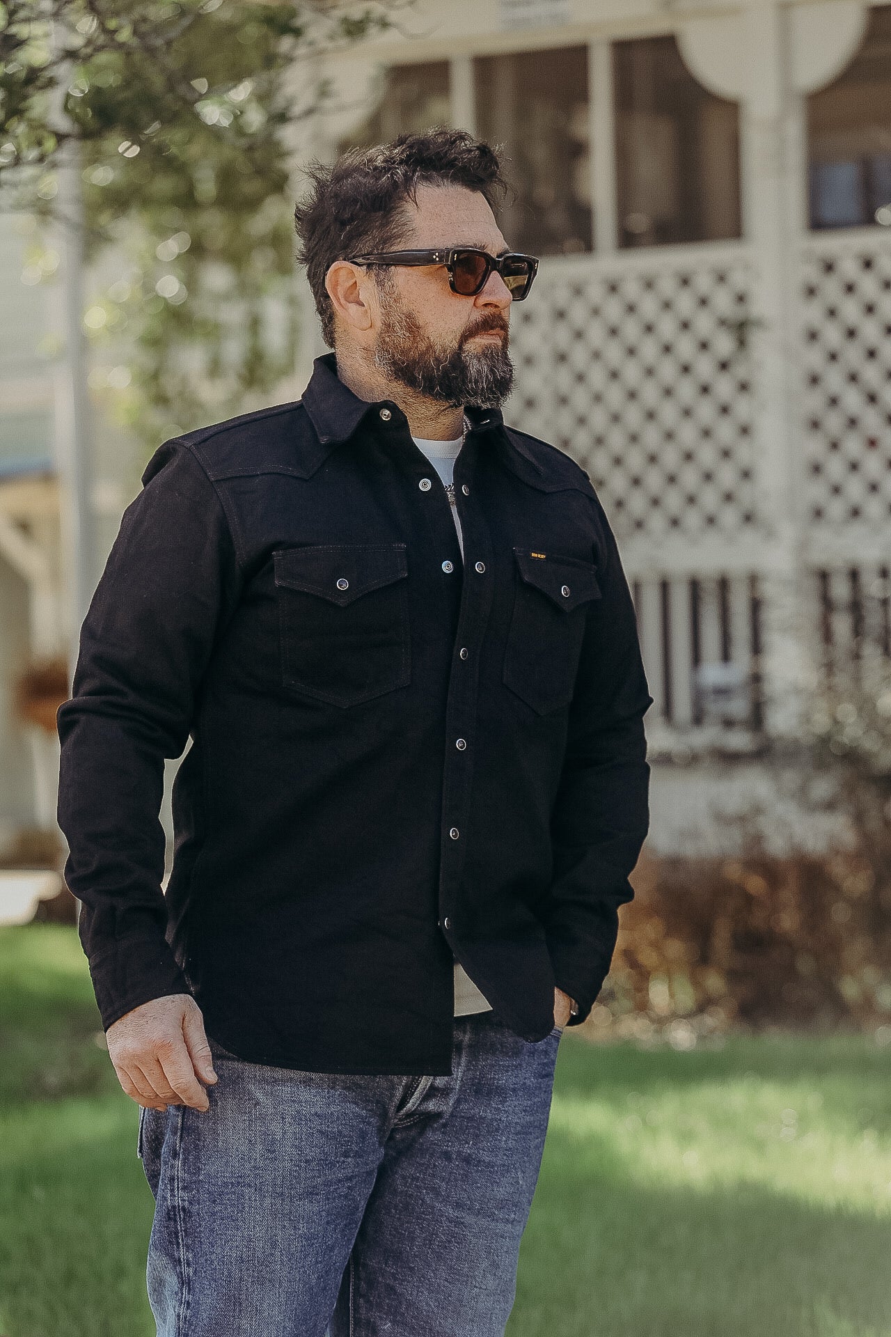 Bearded man wearing sunglasses and black button-up shirt standing outdoors in front of a white house with lattice porch