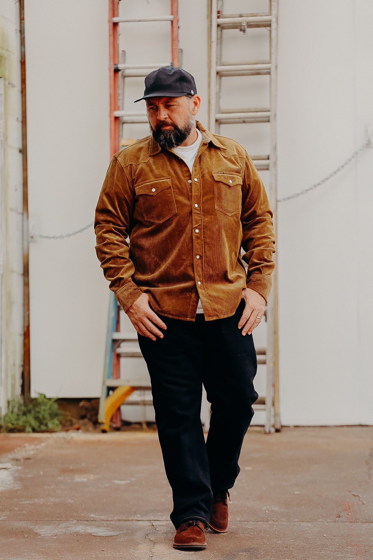A man wearing a brown corduroy western shirt with a collar, button-up front, and cap, standing in front of a white background with a ladder in the background.