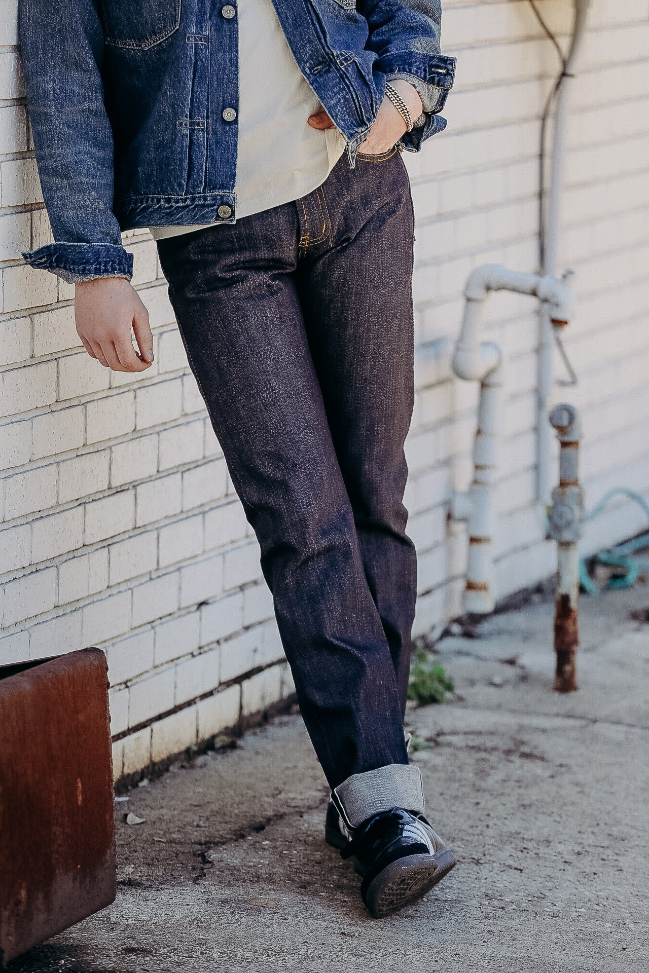 Person wearing dark denim jeans cuffed at ankle, black sneakers, denim jacket, standing by white brick wall