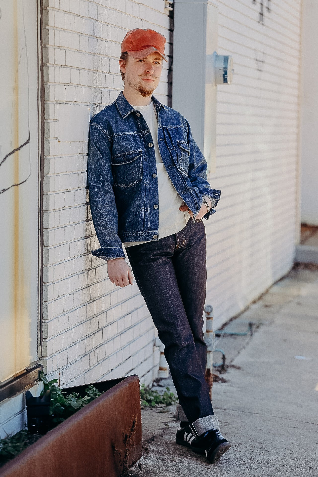 Man wearing denim jacket, dark jeans, white shirt, orange cap, and black sneakers leaning on a white brick wall